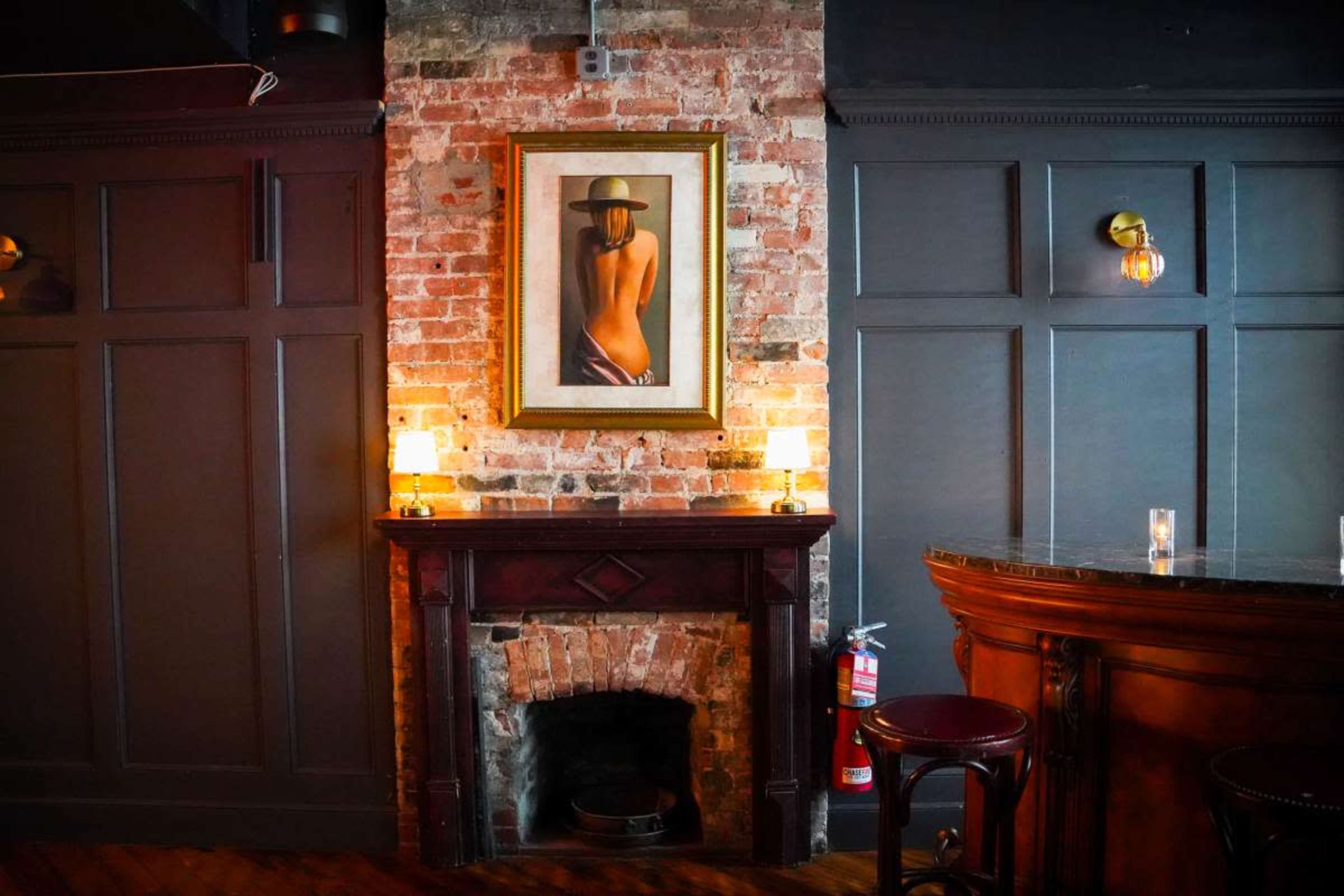 The image shows a restaurant or bar interior featuring a brick wall with a framed painting of a woman, a dark wooden mantelpiece, and a bar counter with low lighting.