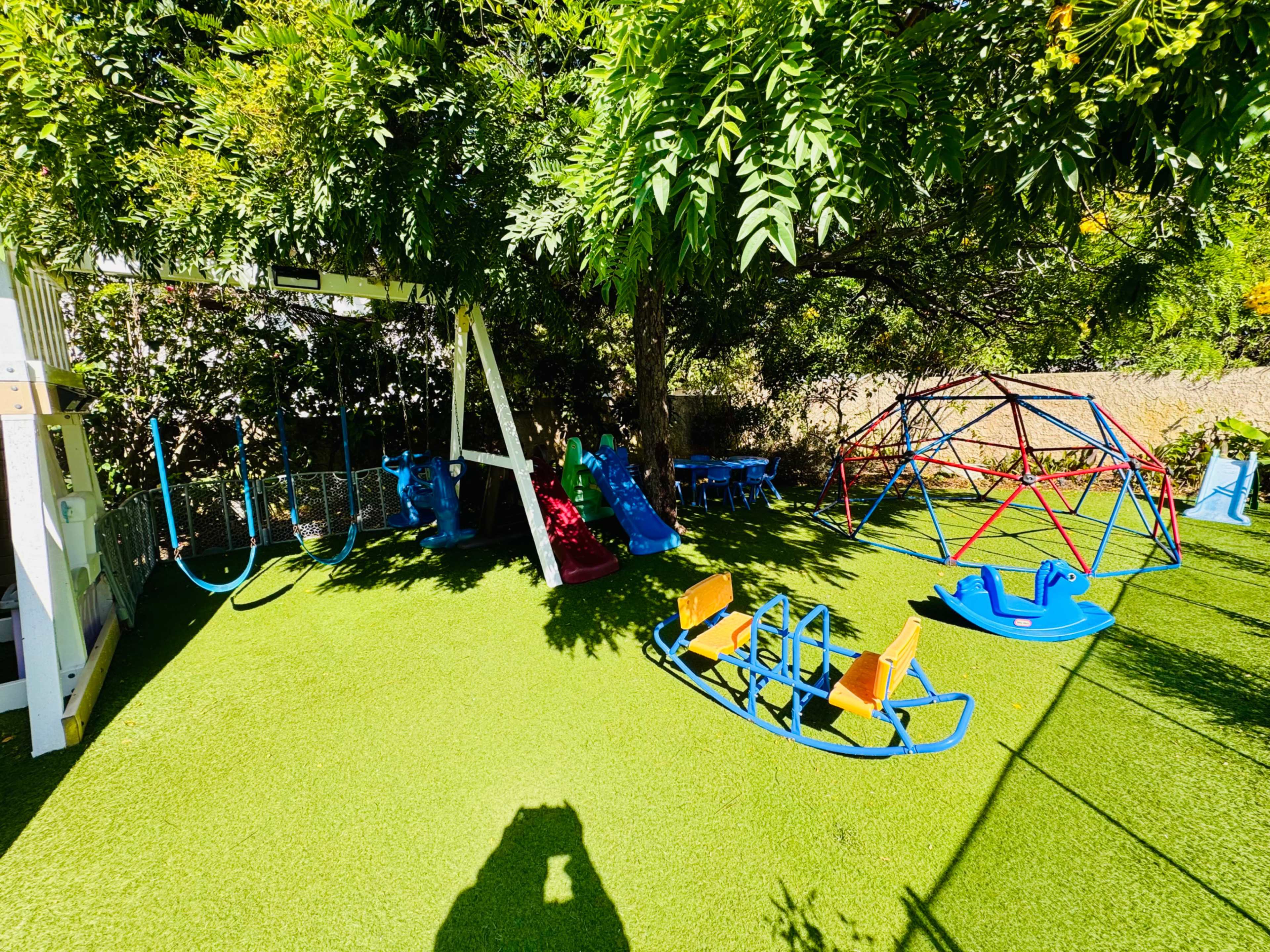 The scene shows a colorful children's play area featuring swings, slides, a climbing dome, and a seesaw, all situated on a grassy surface under a shaded tree.