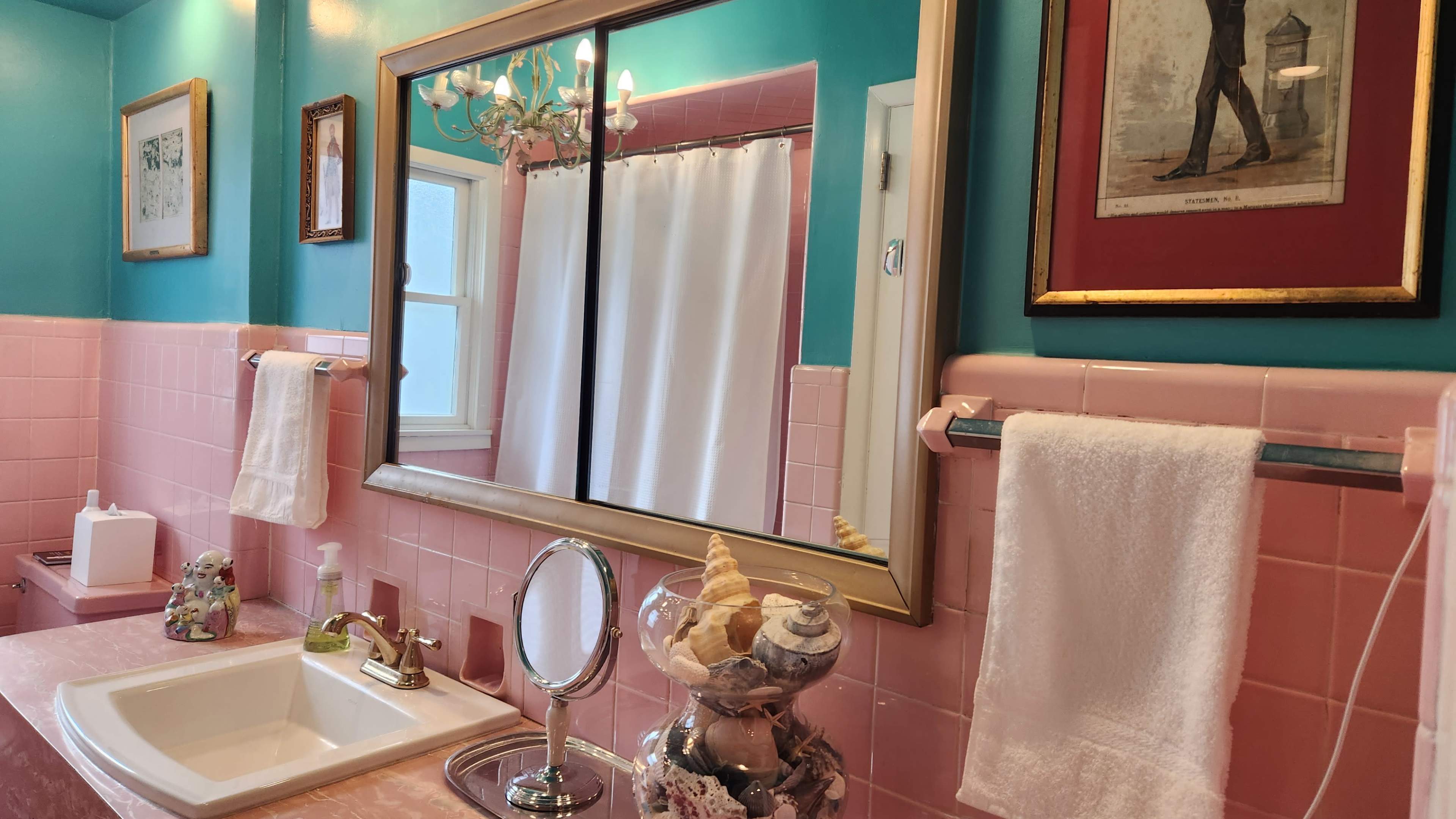 A retro-style bathroom features pink tiles and a large mirror, with a decorative chandelier and seashells displayed in a glass container on the sink.