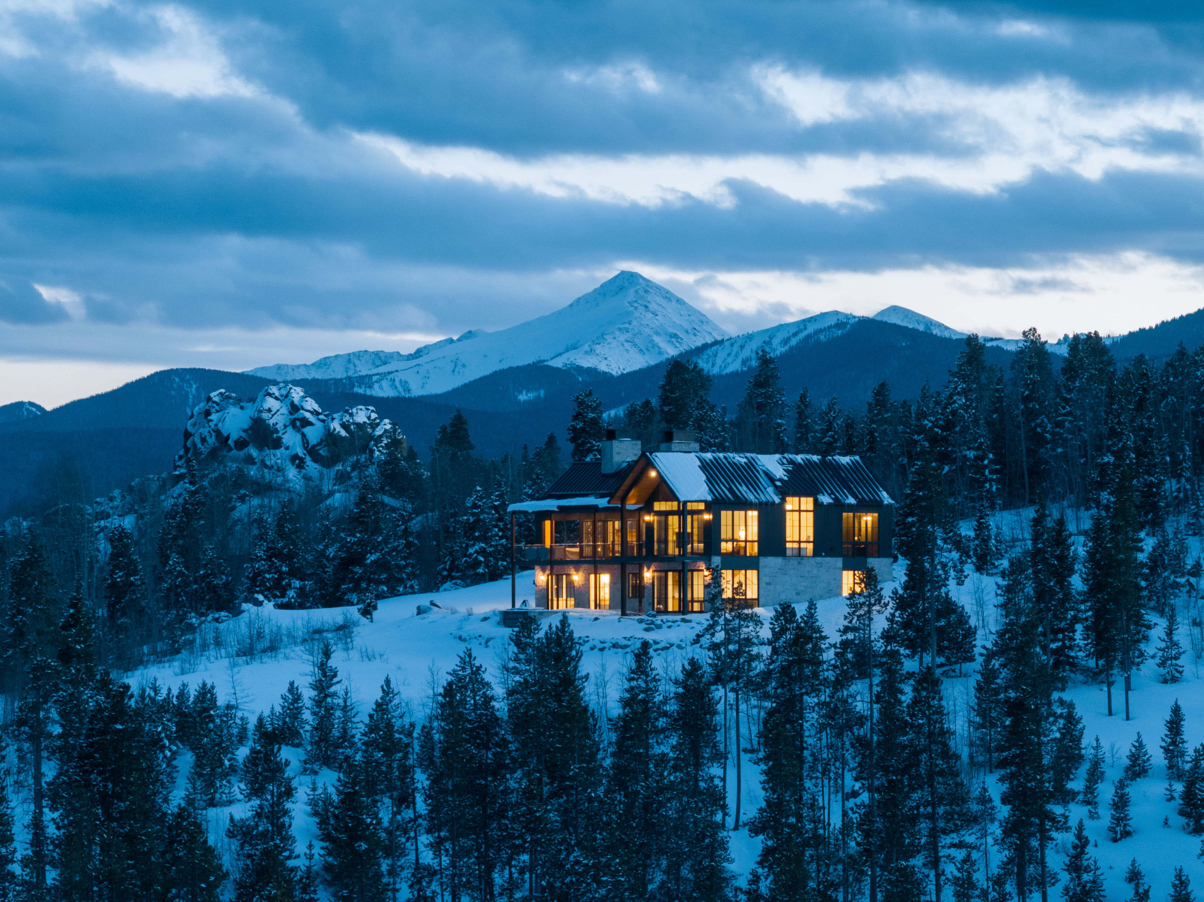 A modern mountain house with illuminated windows is nestled among snow-covered trees, with a backdrop of peaks under a cloudy sky.