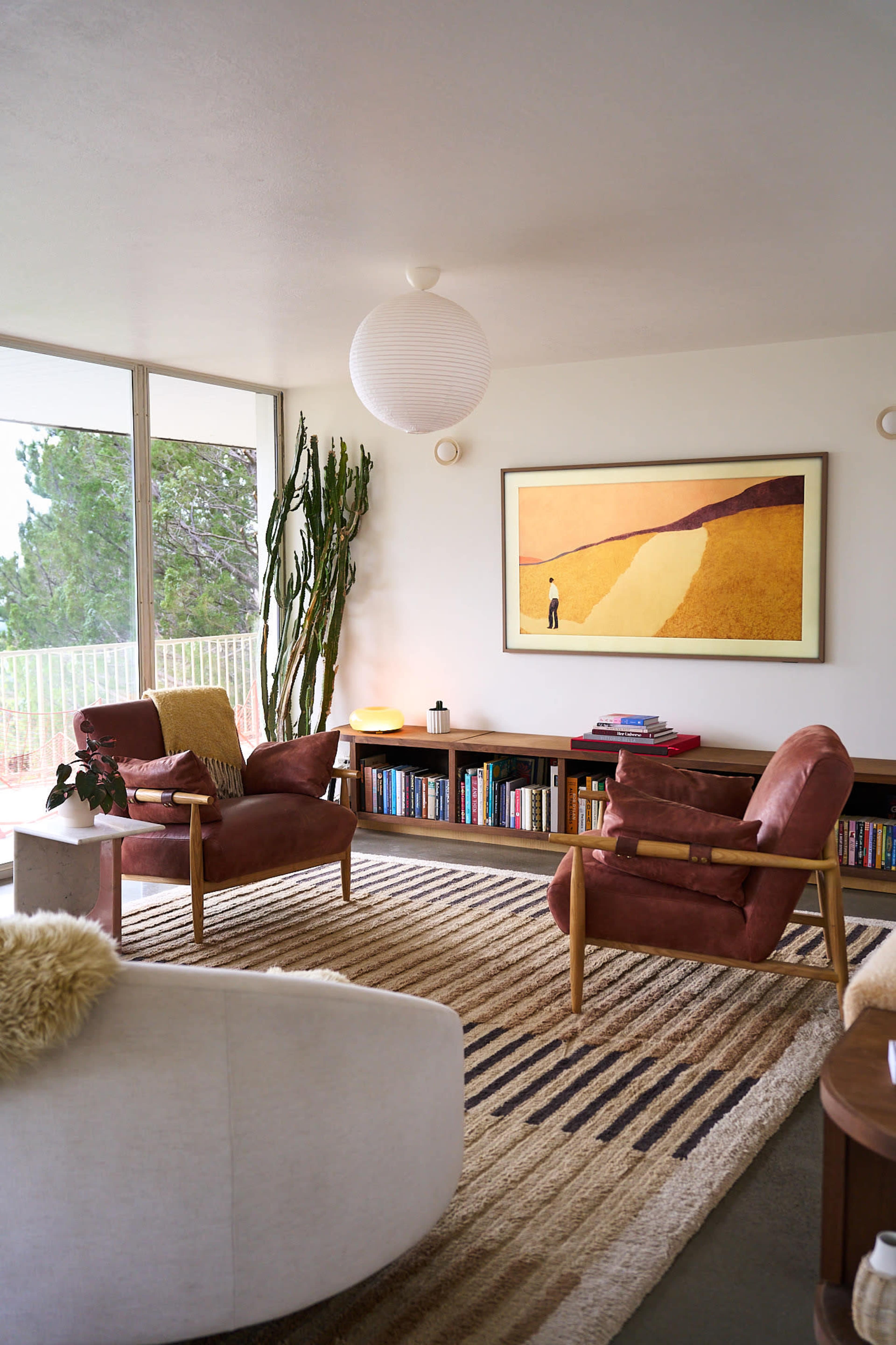 The image shows a modern living room with two brown armchairs, a large abstract painting on the wall, a bookshelf filled with books, and a rug on the floor.