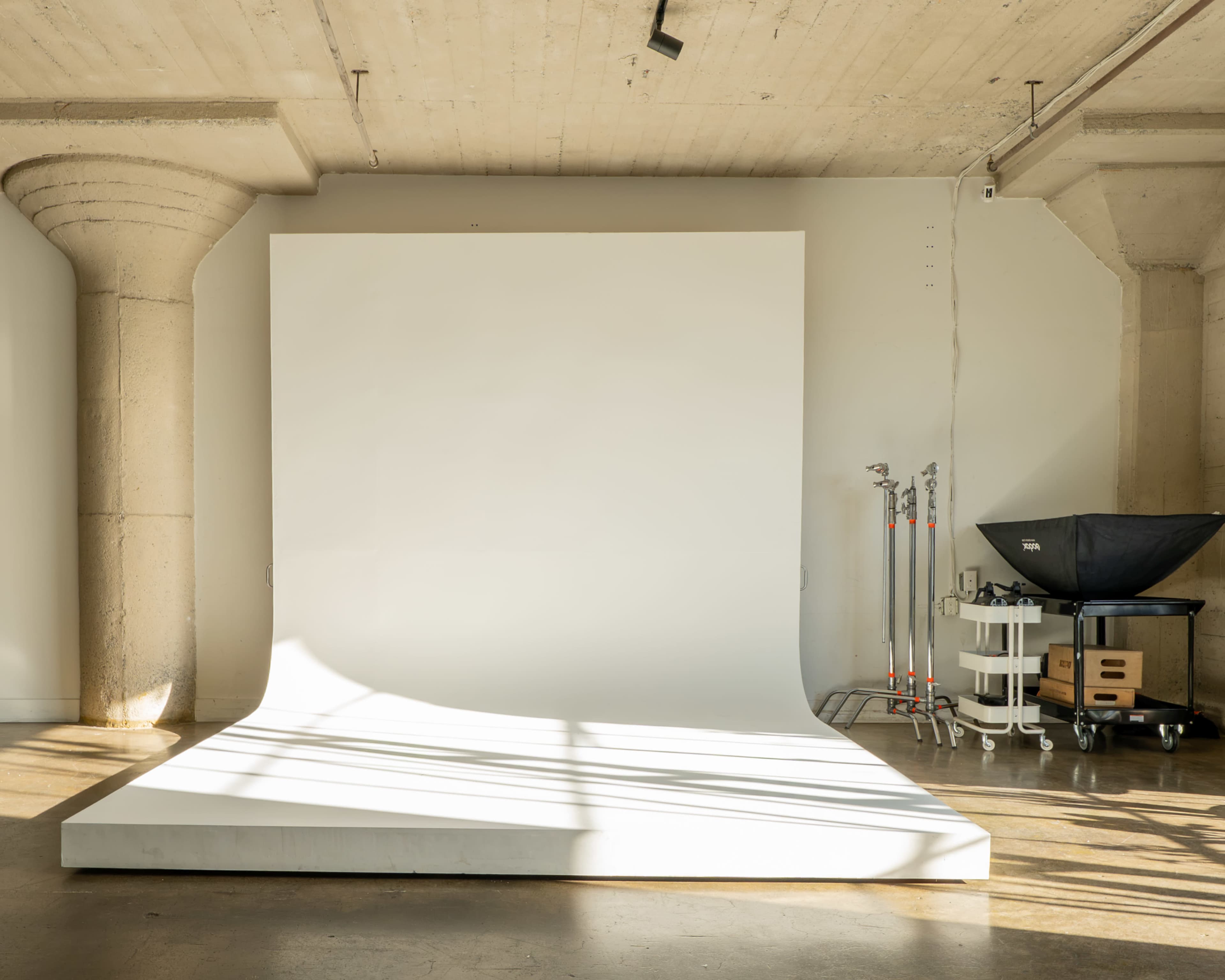 The image shows a minimalist photography studio featuring a curved white backdrop, soft lighting, and equipment neatly arranged along one wall.