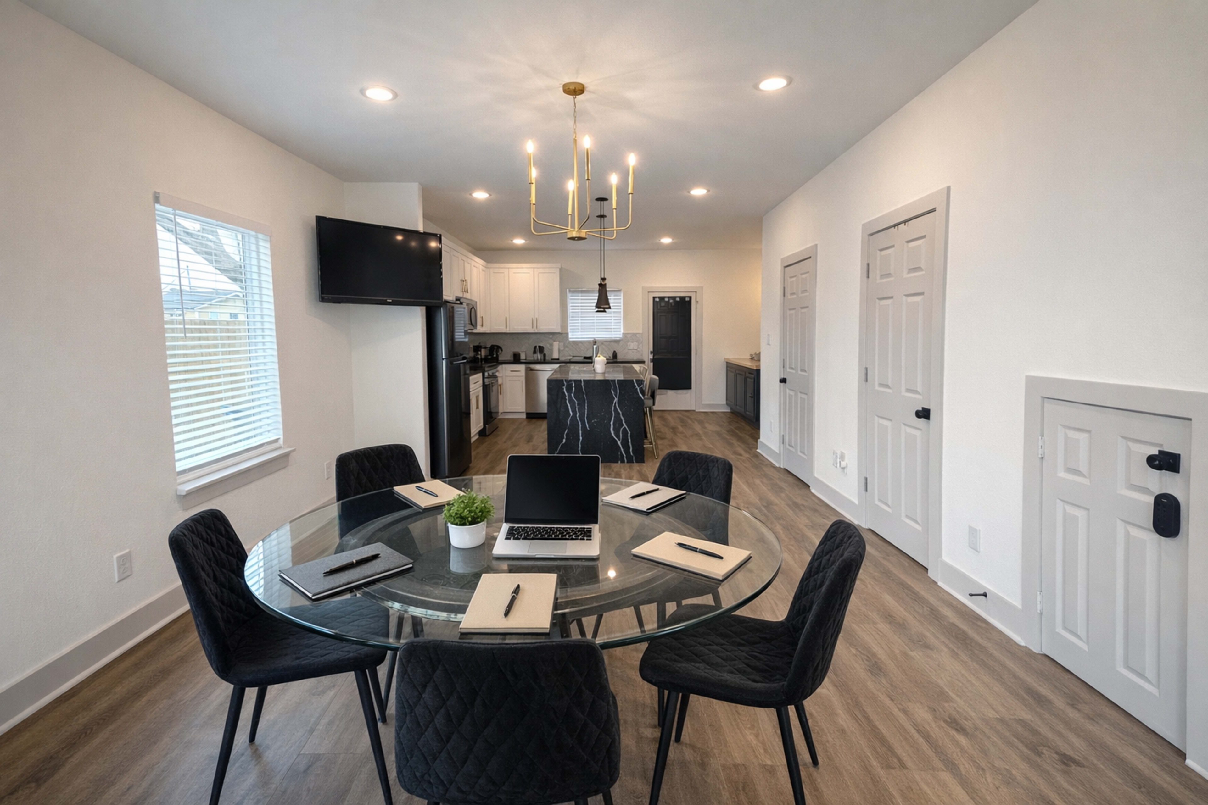 The image shows a modern conference room with a round glass table surrounded by black chairs, a laptop, and notebooks placed on a wooden floor, leading into a kitchen area.