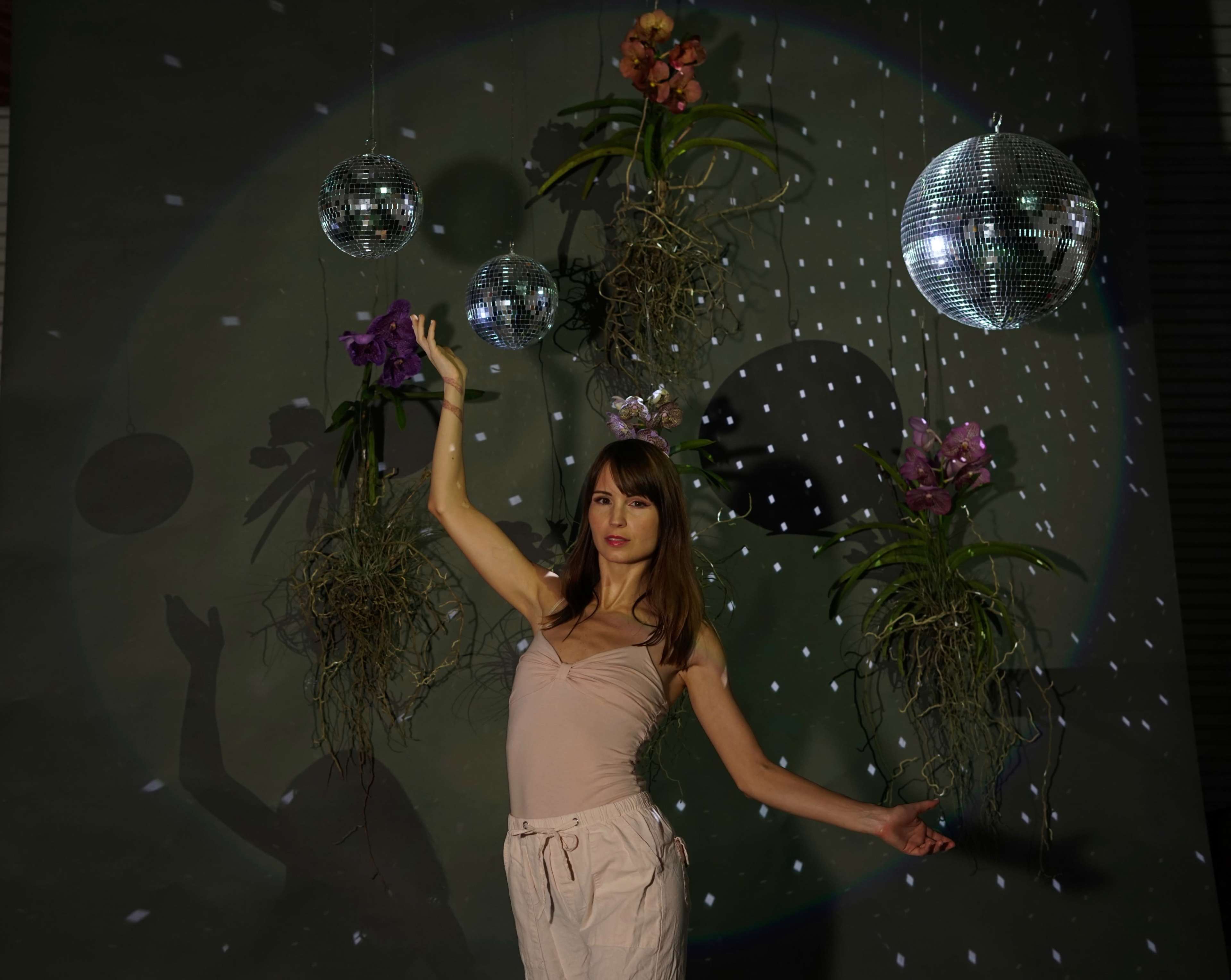 A woman in a light-colored outfit poses with one arm raised in front of disco balls and hanging plants against a dark background.