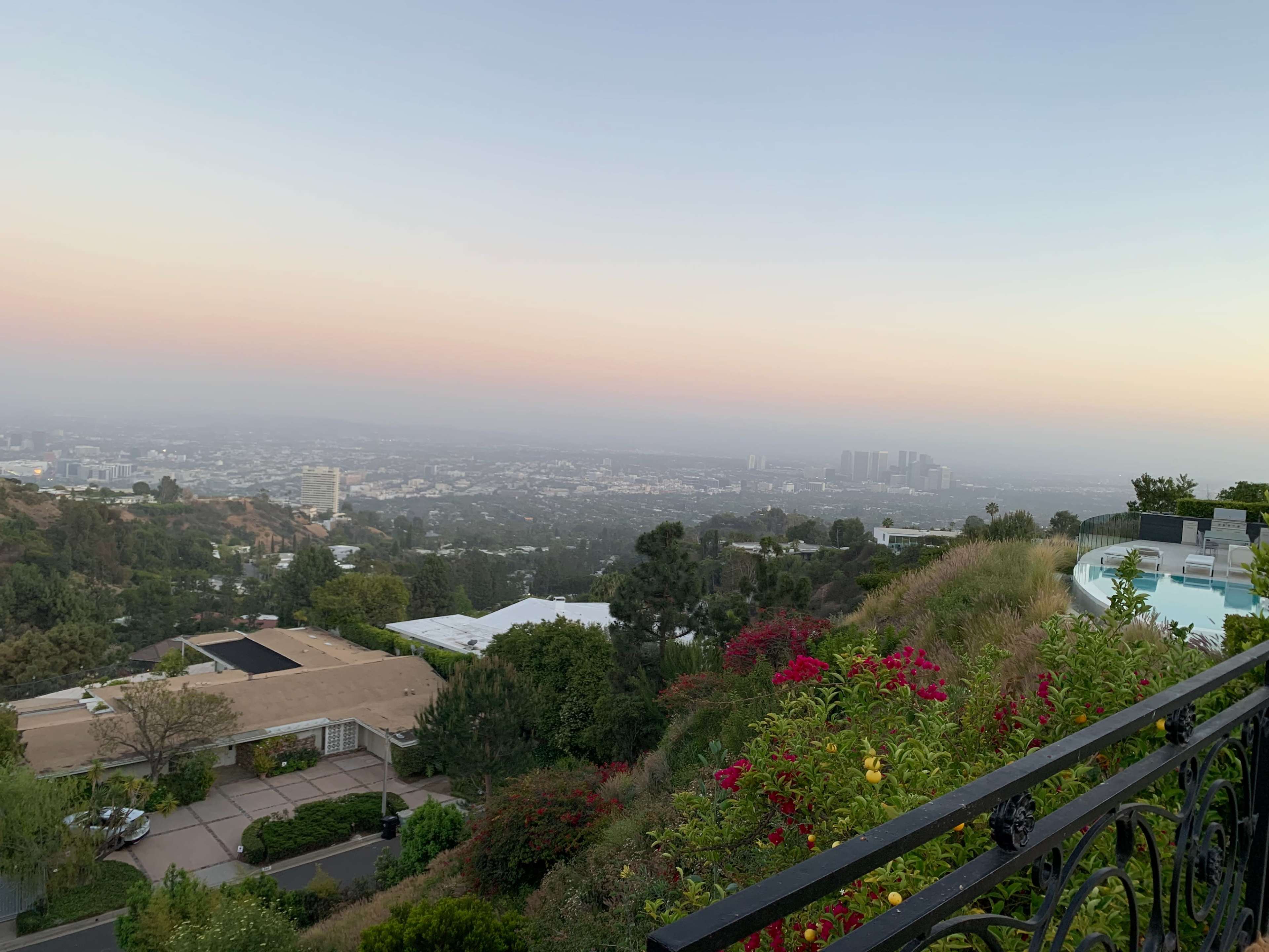 The image features a hillside view overlooking a cityscape at dusk, with houses and a swimming pool in the foreground and a hazy skyline in the distance.