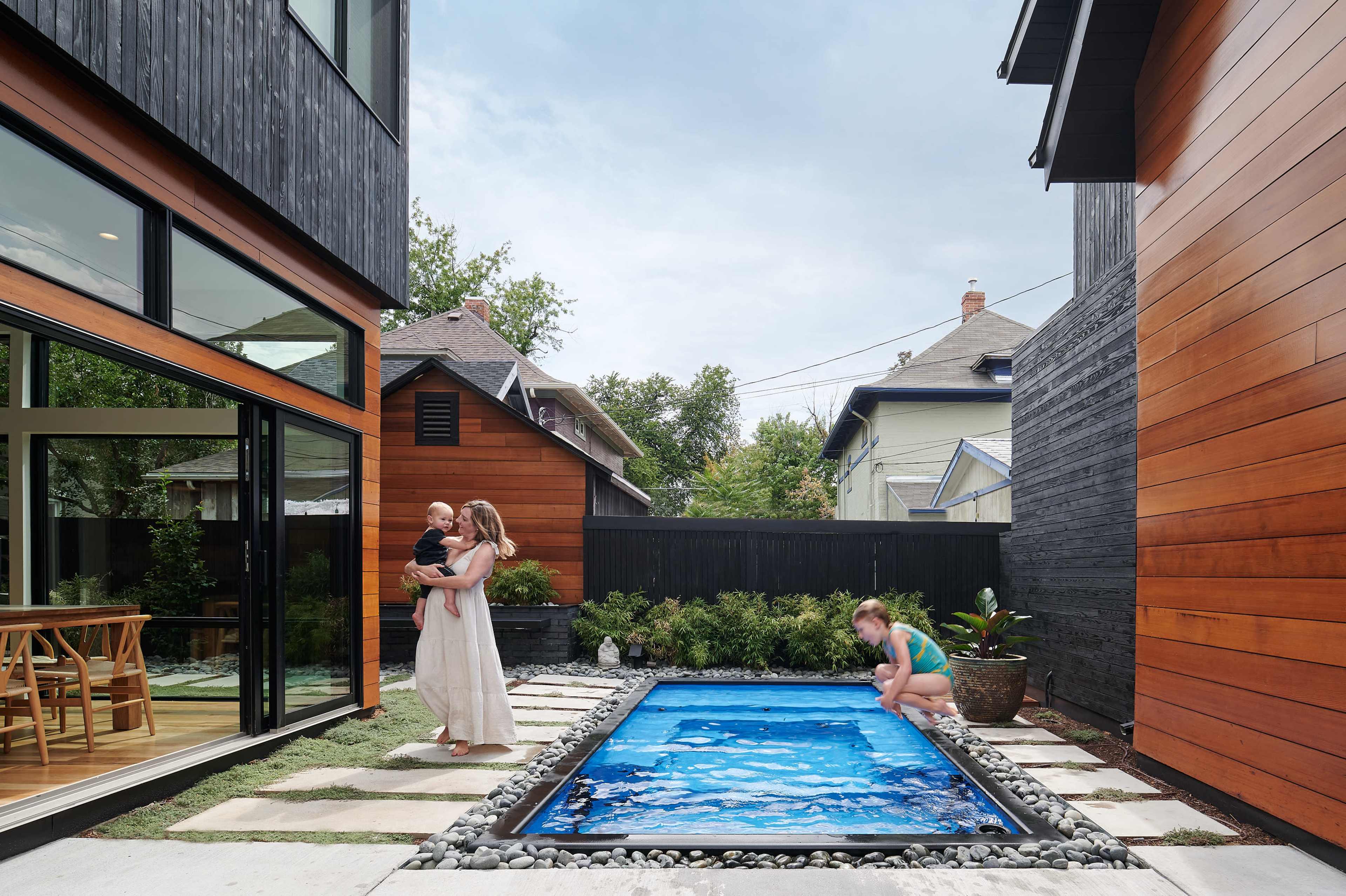 A woman holds a child while standing near a small pool, as a child plays at the edge of the water in a courtyard surrounded by modern houses.