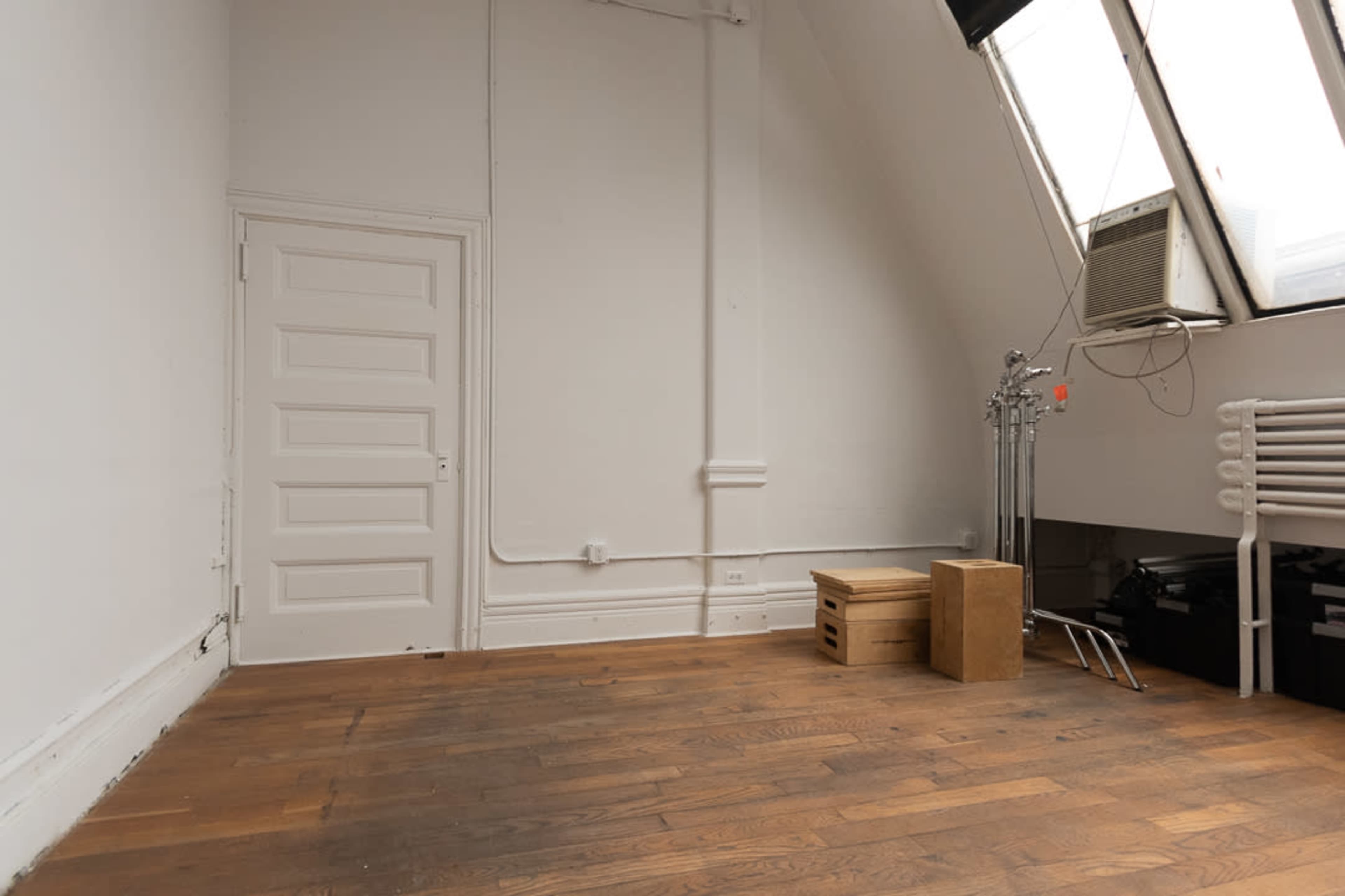 A sparsely furnished room with a wooden floor, a white door, an air conditioning unit on the wall, and a few boxes.