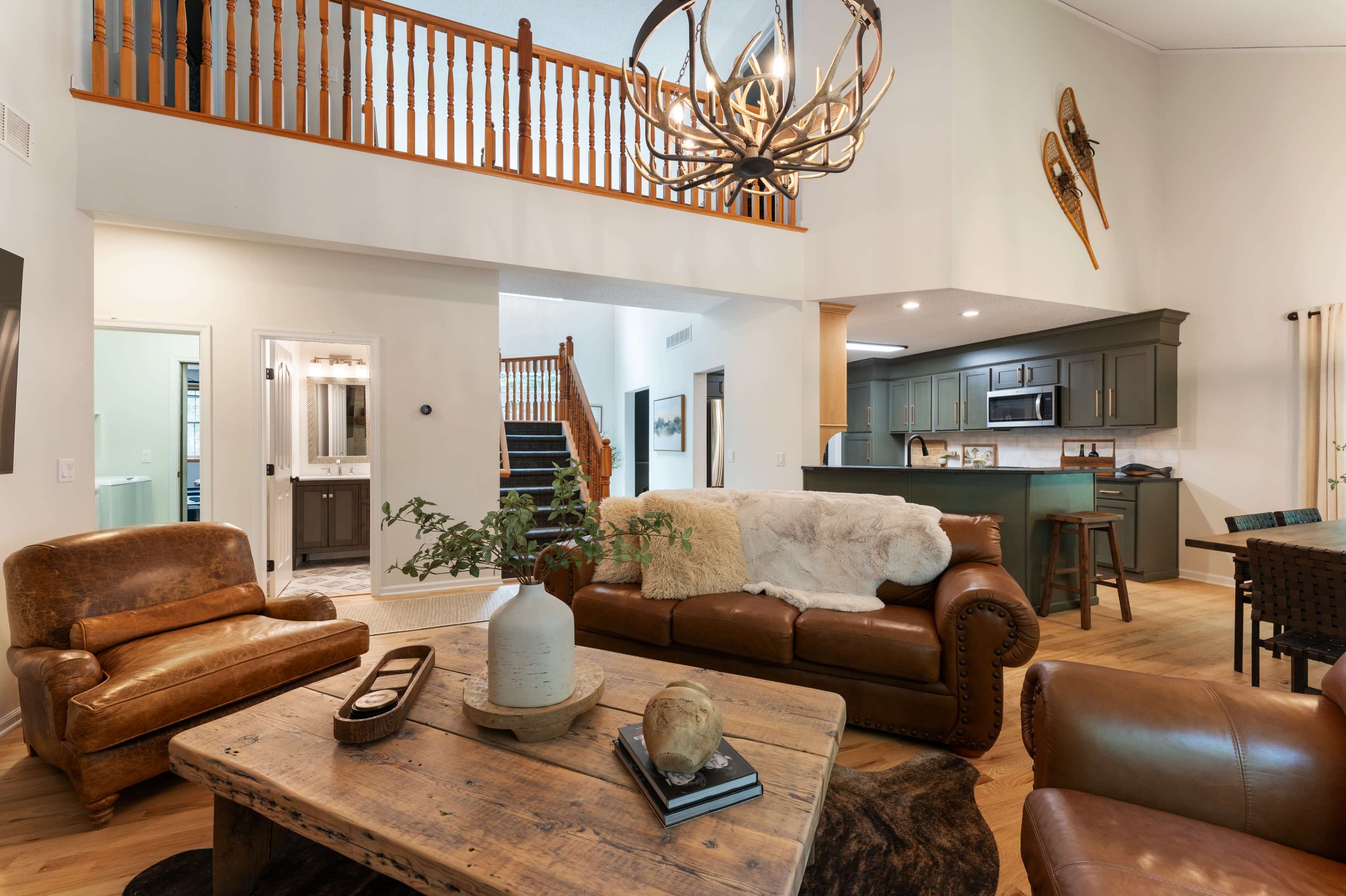 The image shows a spacious living area with a high ceiling, featuring leather sofas, a large wooden coffee table, and an open view of a kitchen and staircase.