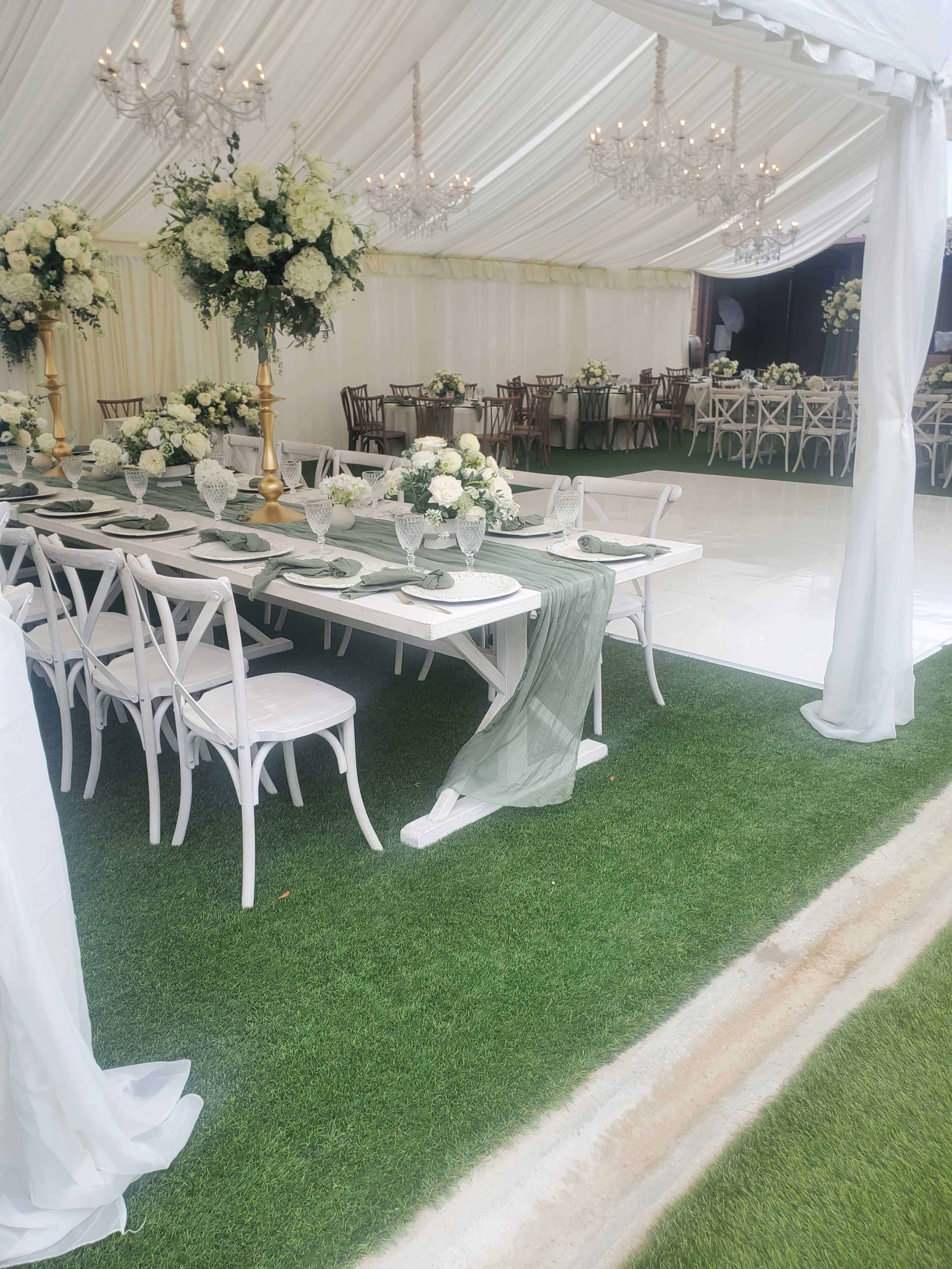A tented event space features long tables set with floral centerpieces and elegant dinnerware, surrounded by chairs and decorative chandeliers.