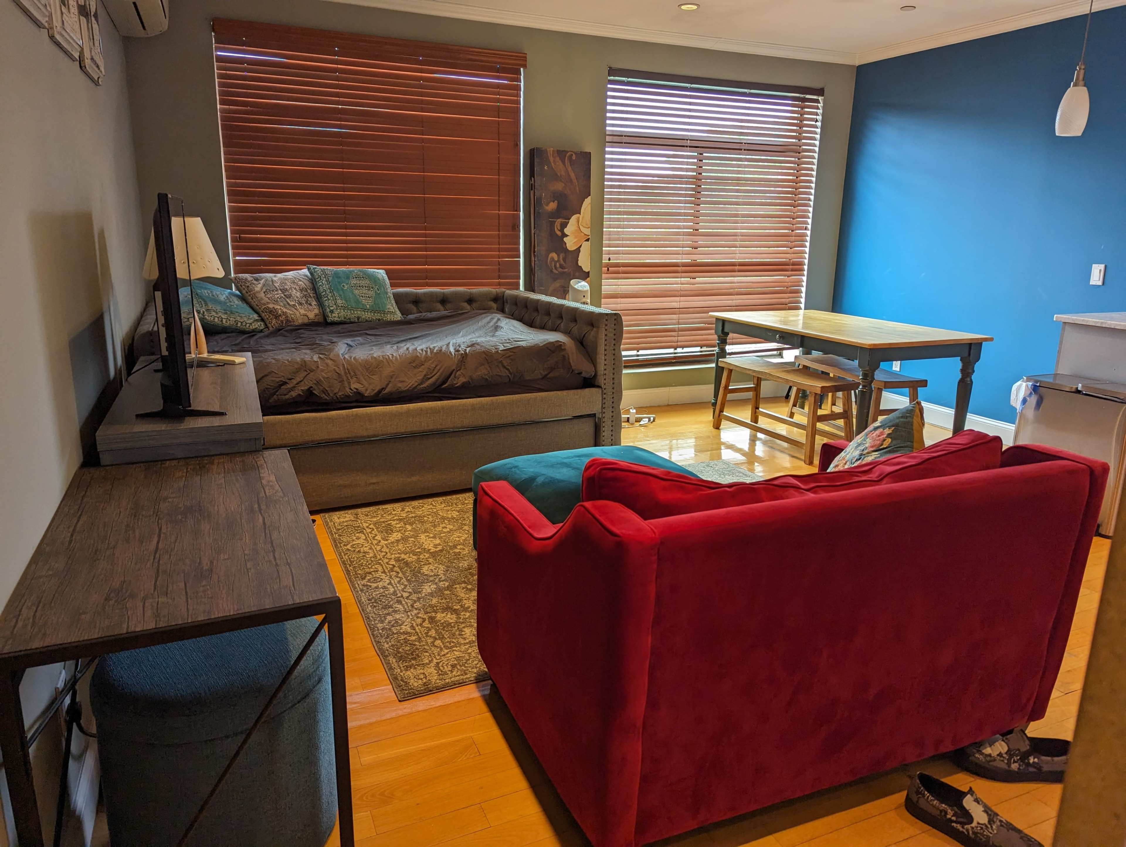 The image shows a well-furnished living space with a daybed, a red sofa, a wooden dining table, and large windows with blinds.