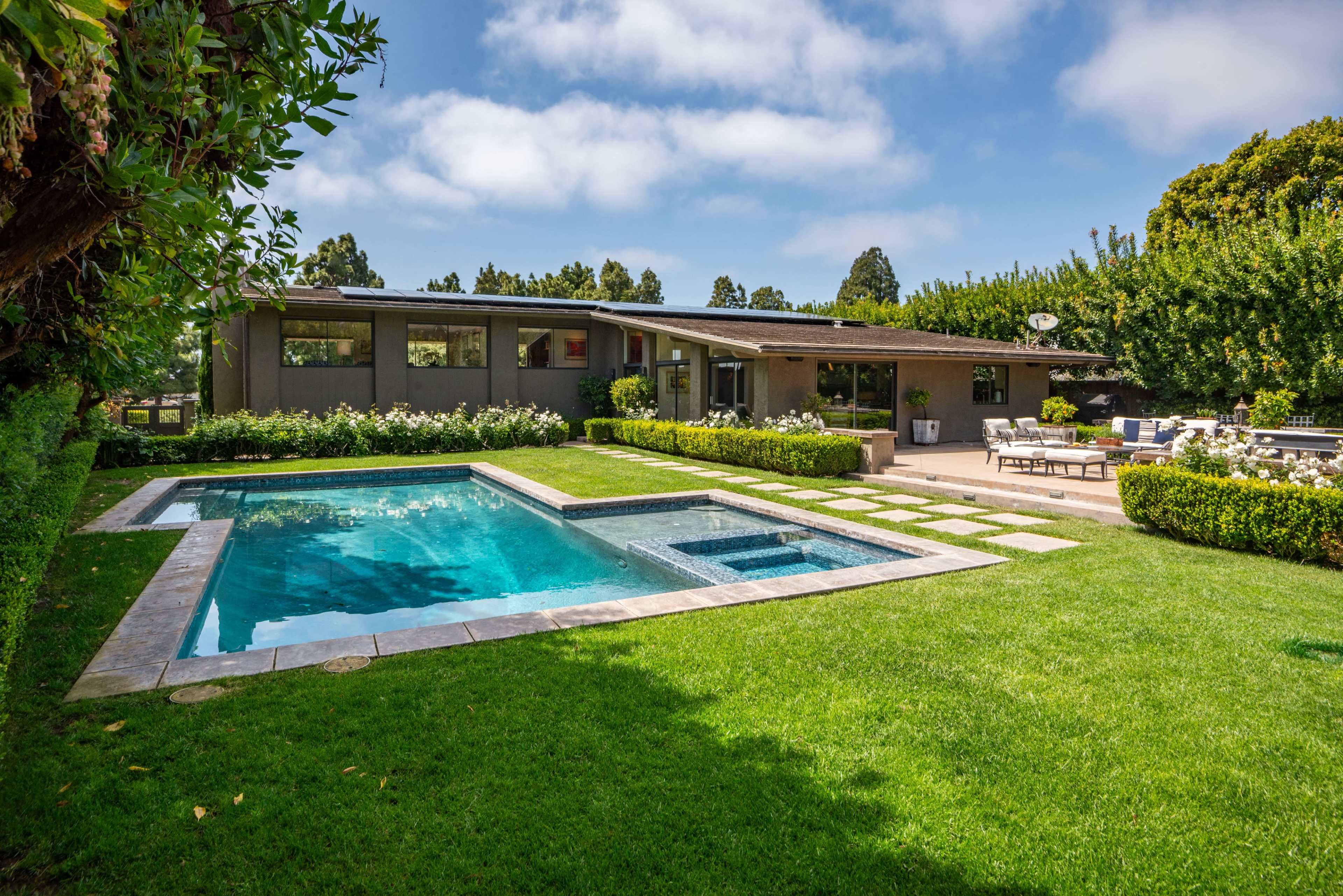 The image shows a modern house with a backyard featuring a swimming pool, spa, and landscaped lawn with neatly arranged stone pathways and seating areas.