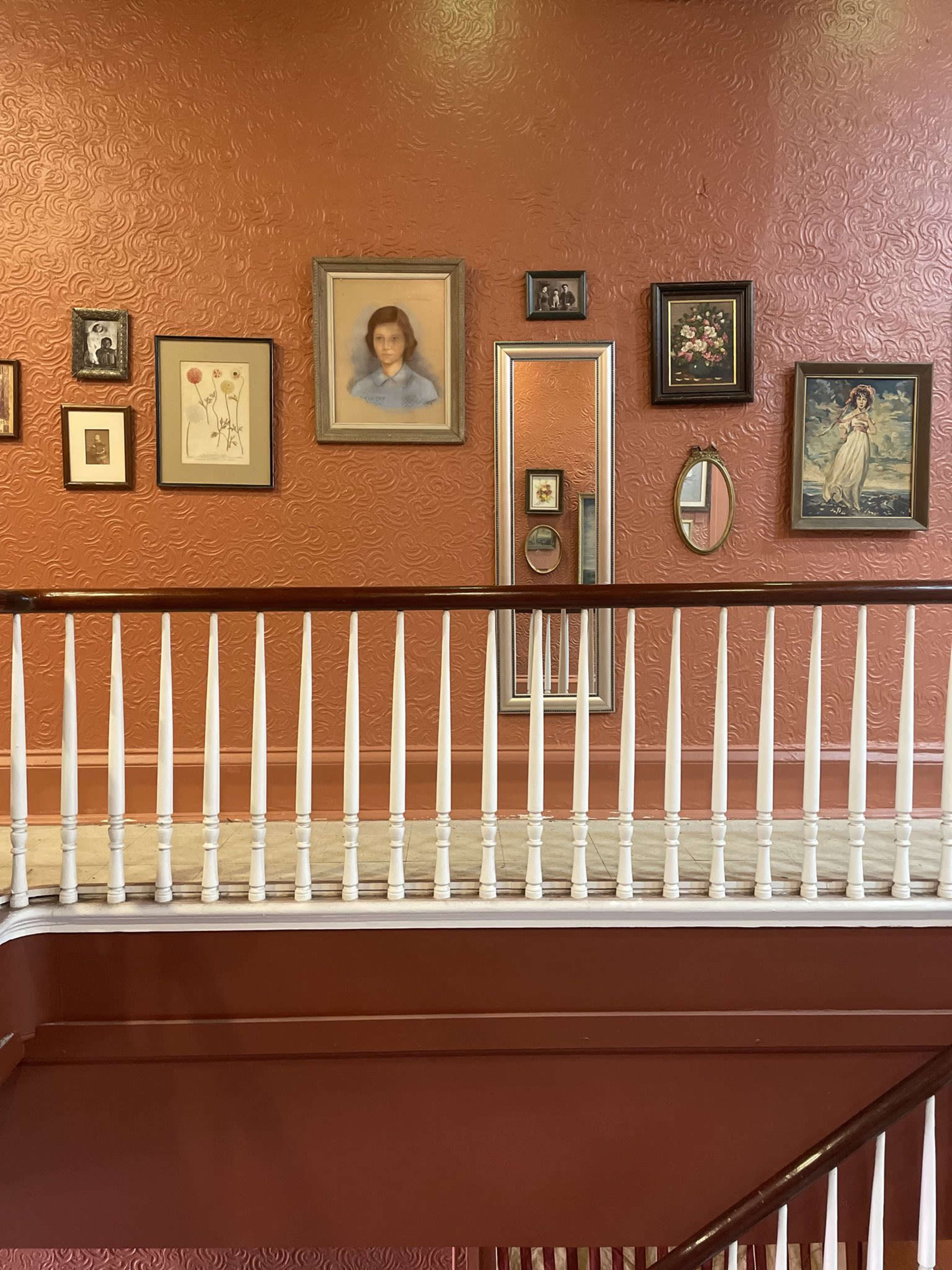 The image shows an interior staircase with a railing, featuring a wall adorned with various framed pictures and a mirror against a textured orange background.