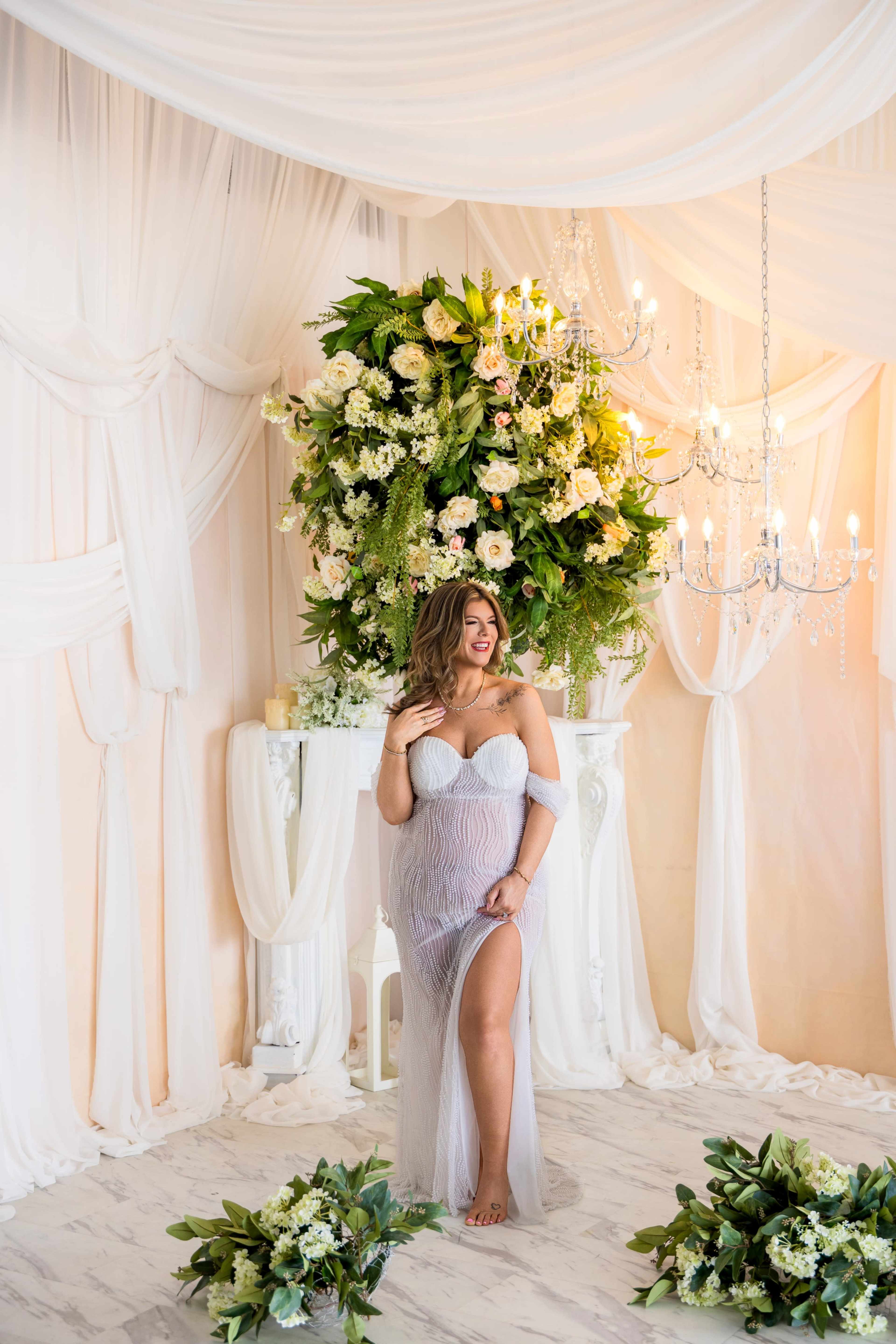 A woman poses in a light-colored gown against a backdrop of draped fabric and floral arrangements.