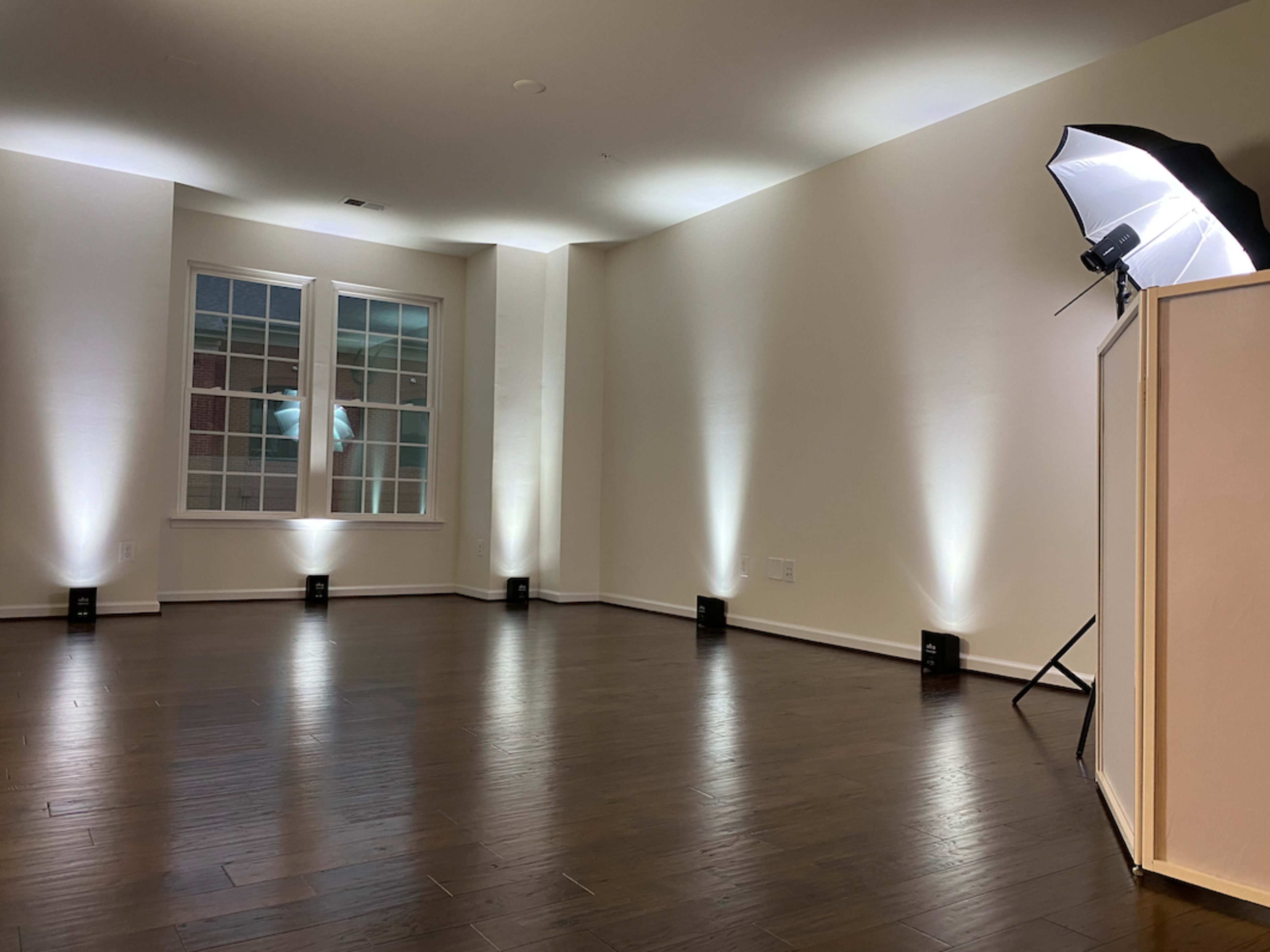 A spacious room with wooden flooring and soft lighting emanating from wall-mounted fixtures and a large umbrella softbox in the corner.