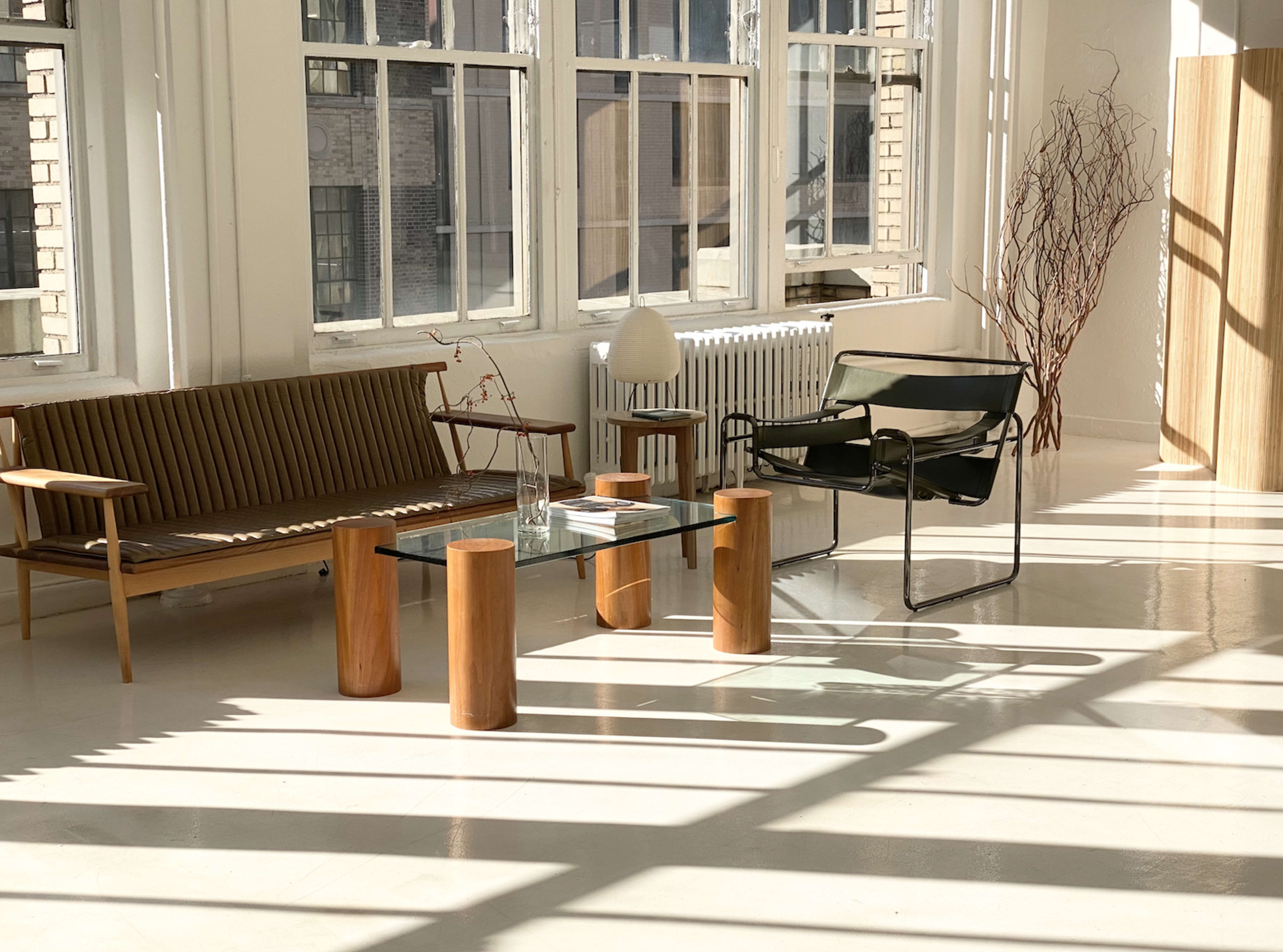 The scene features a bright, minimalist room with a wooden sofa, a glass coffee table supported by cylindrical wooden legs, and a green chair beside a tall wooden structure.