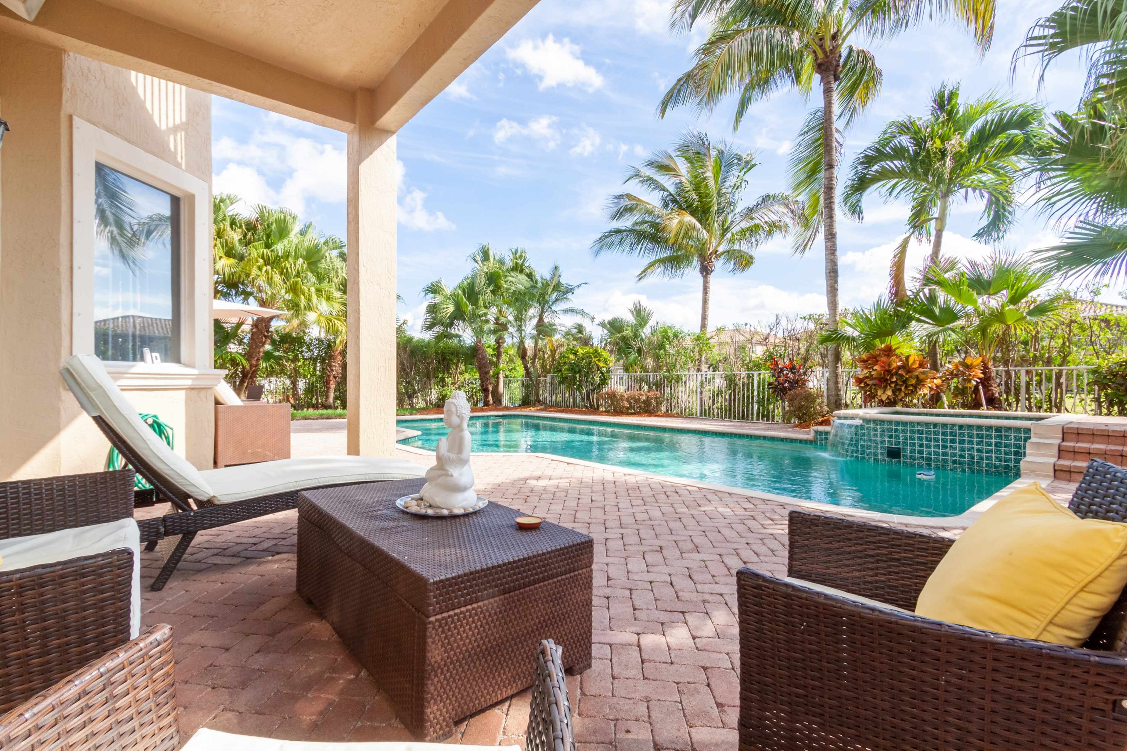 A patio area features a lounge chair, coffee table, and seating beside a swimming pool surrounded by palm trees.