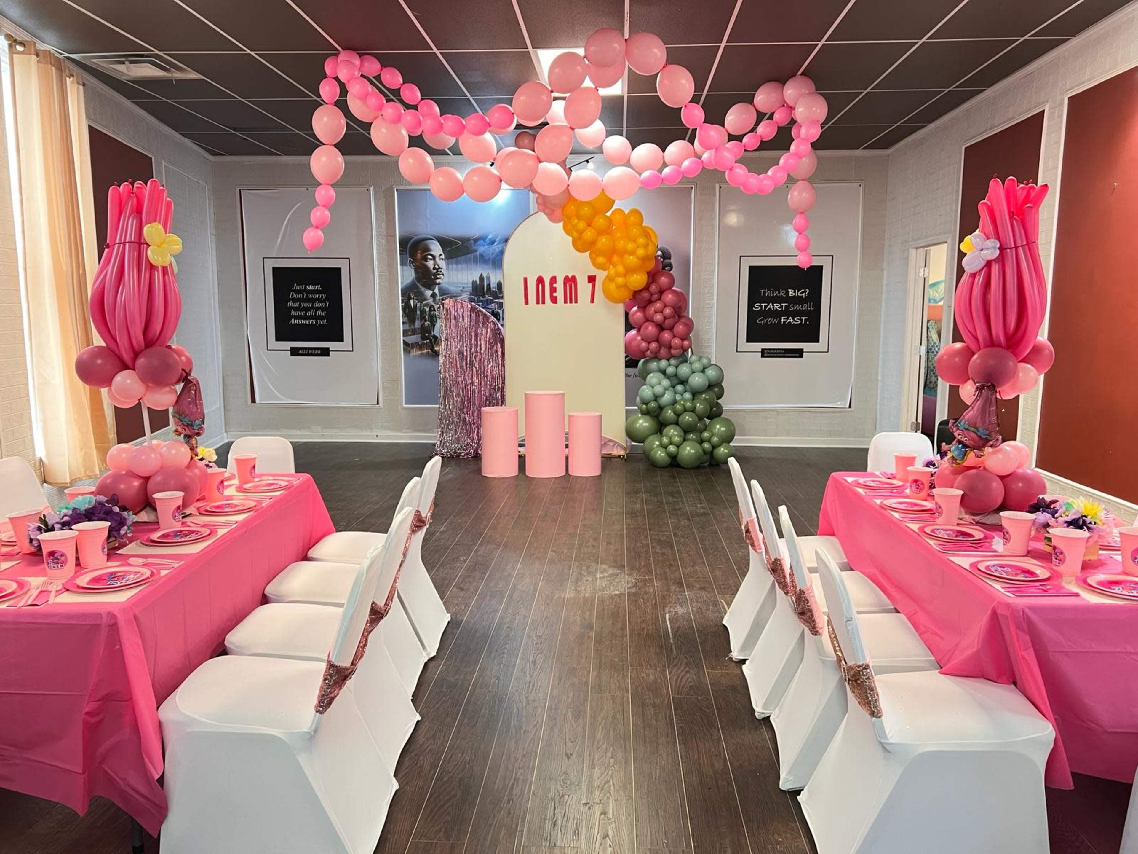 A decorated event space features tables set with pink tableware and balloon arrangements in various colors.