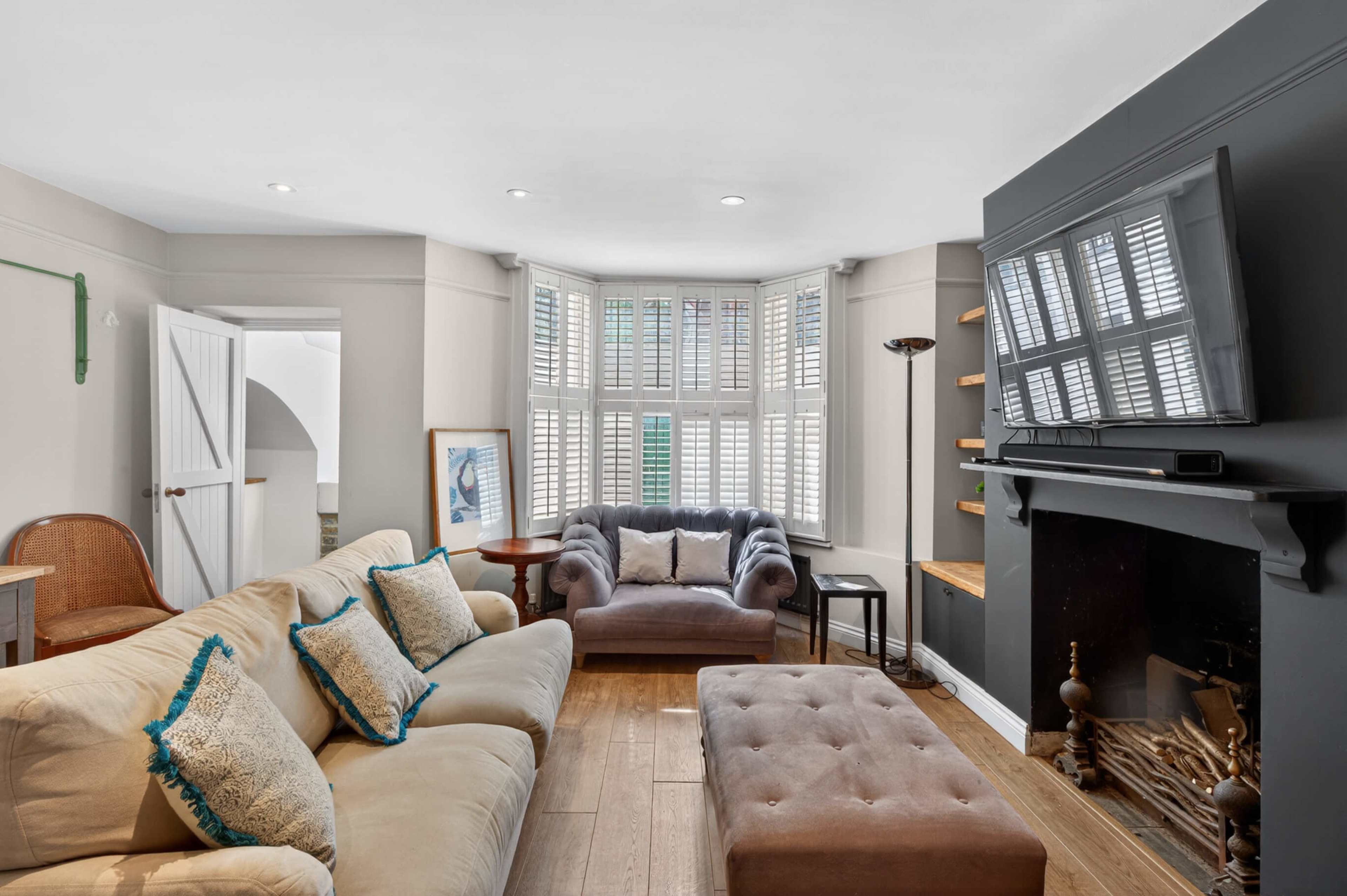 The image shows a cozy living room with a beige sectional sofa, a grey armchair, and a flat-screen TV mounted on a black wall, along with wooden shelves and a fireplace.