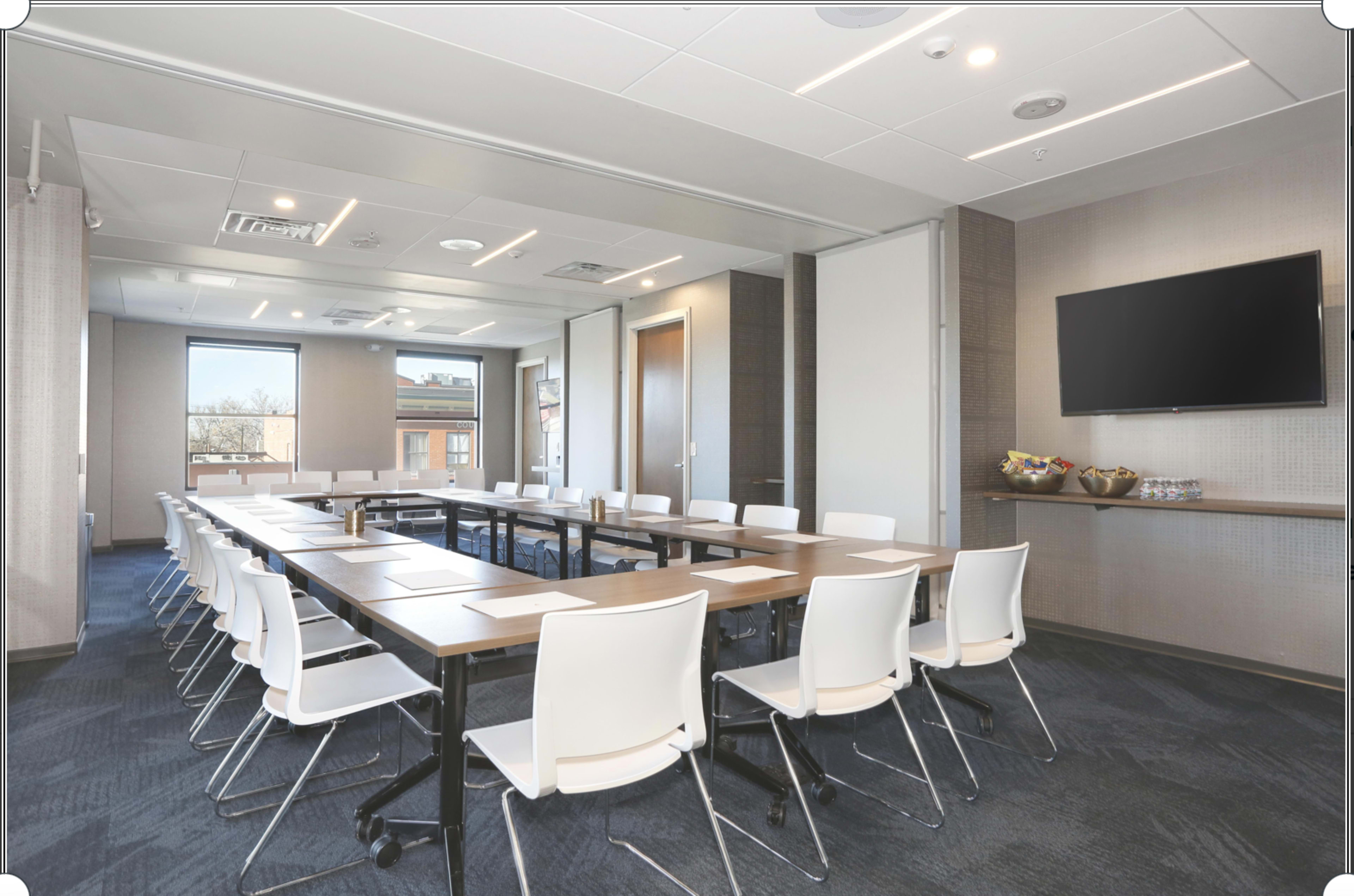 A modern conference room features a long table surrounded by white chairs, with a wall-mounted screen and natural light coming through the windows.