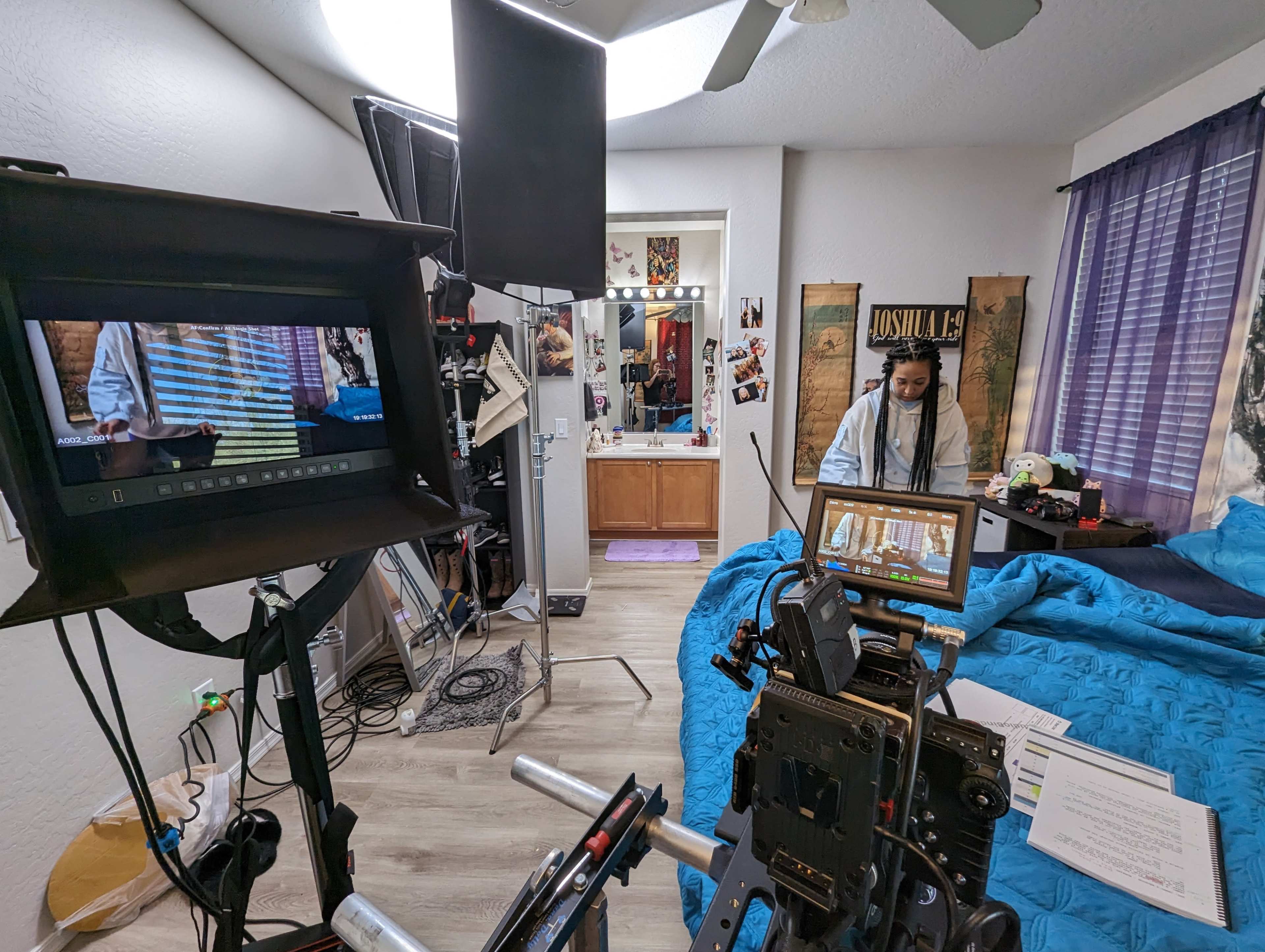 A film set in a bedroom with a camera on a dolly, lighting equipment, and a person in a white outfit interacting with a miniature set.