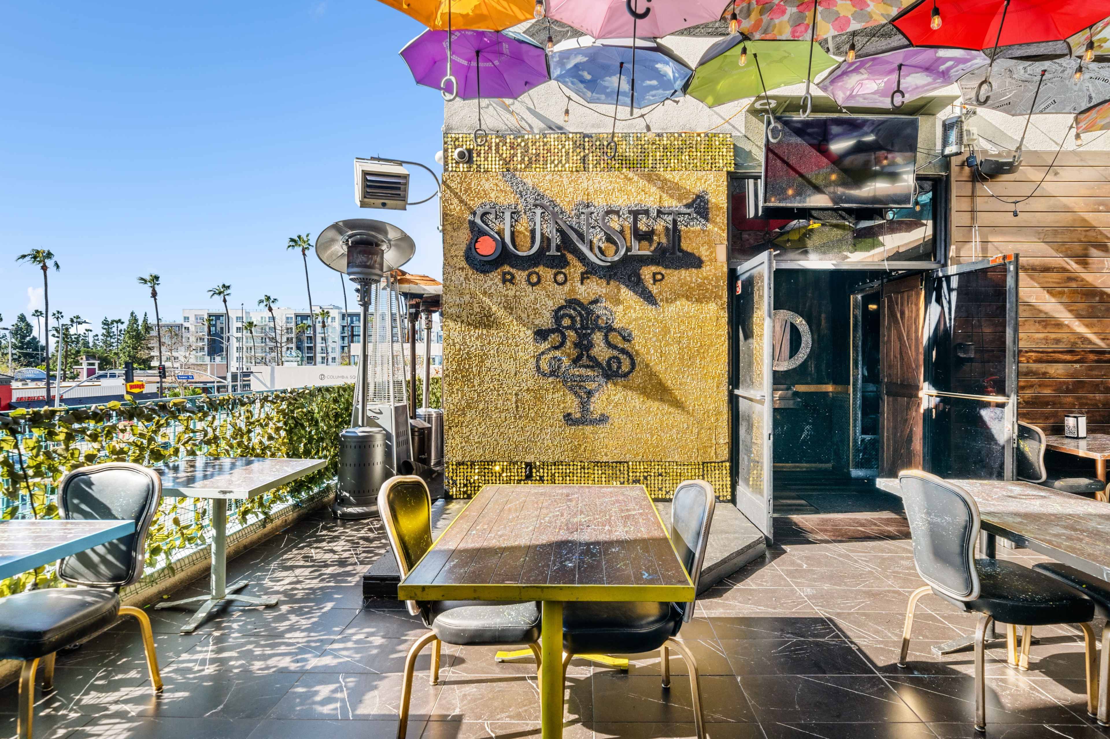 The image shows an outdoor rooftop bar with colorful umbrellas, mosaic wall art, and tables set for customers.