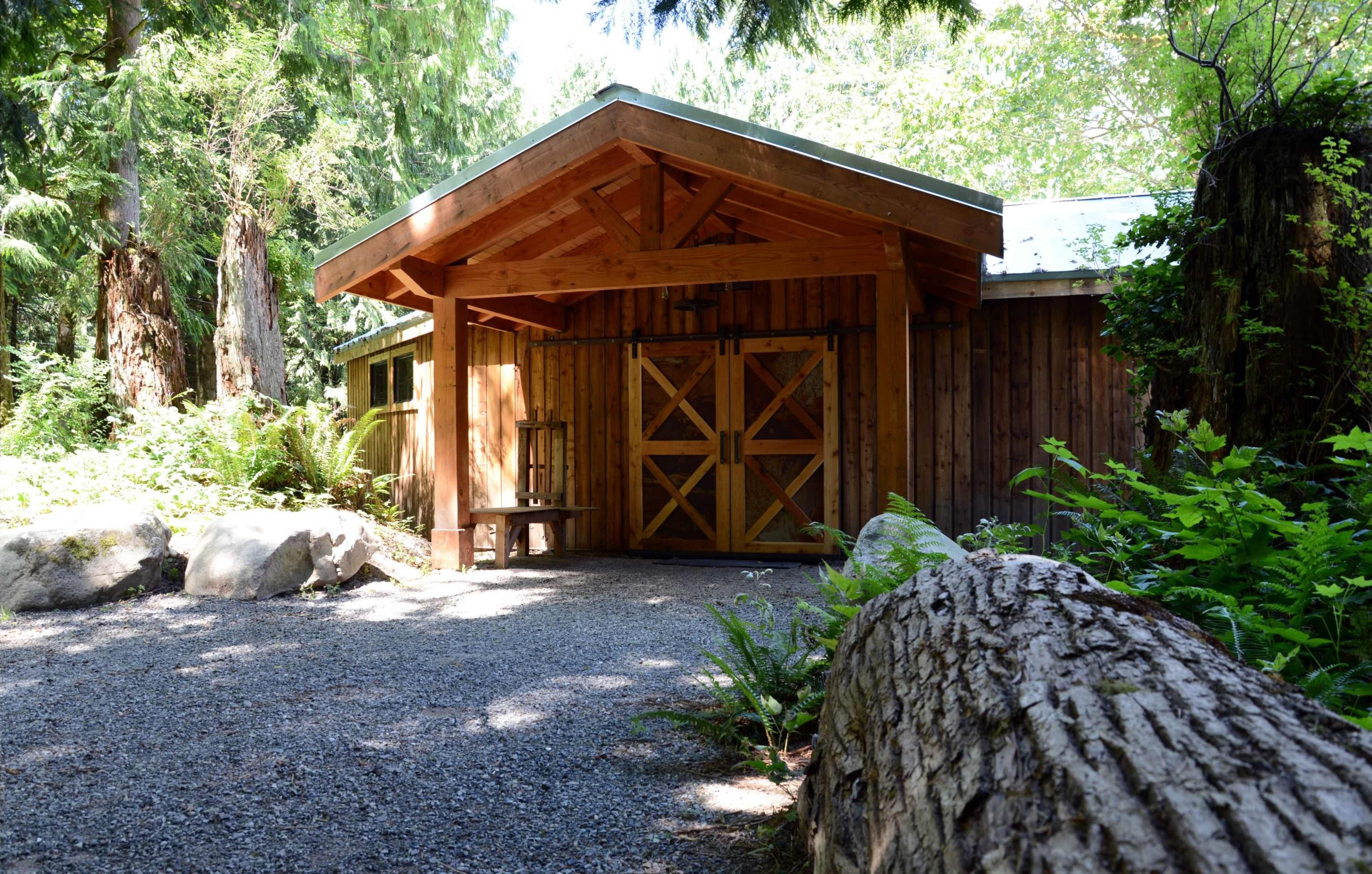 A wooden structure with a peaked roof and double doors is surrounded by trees and ferns along a gravel path.