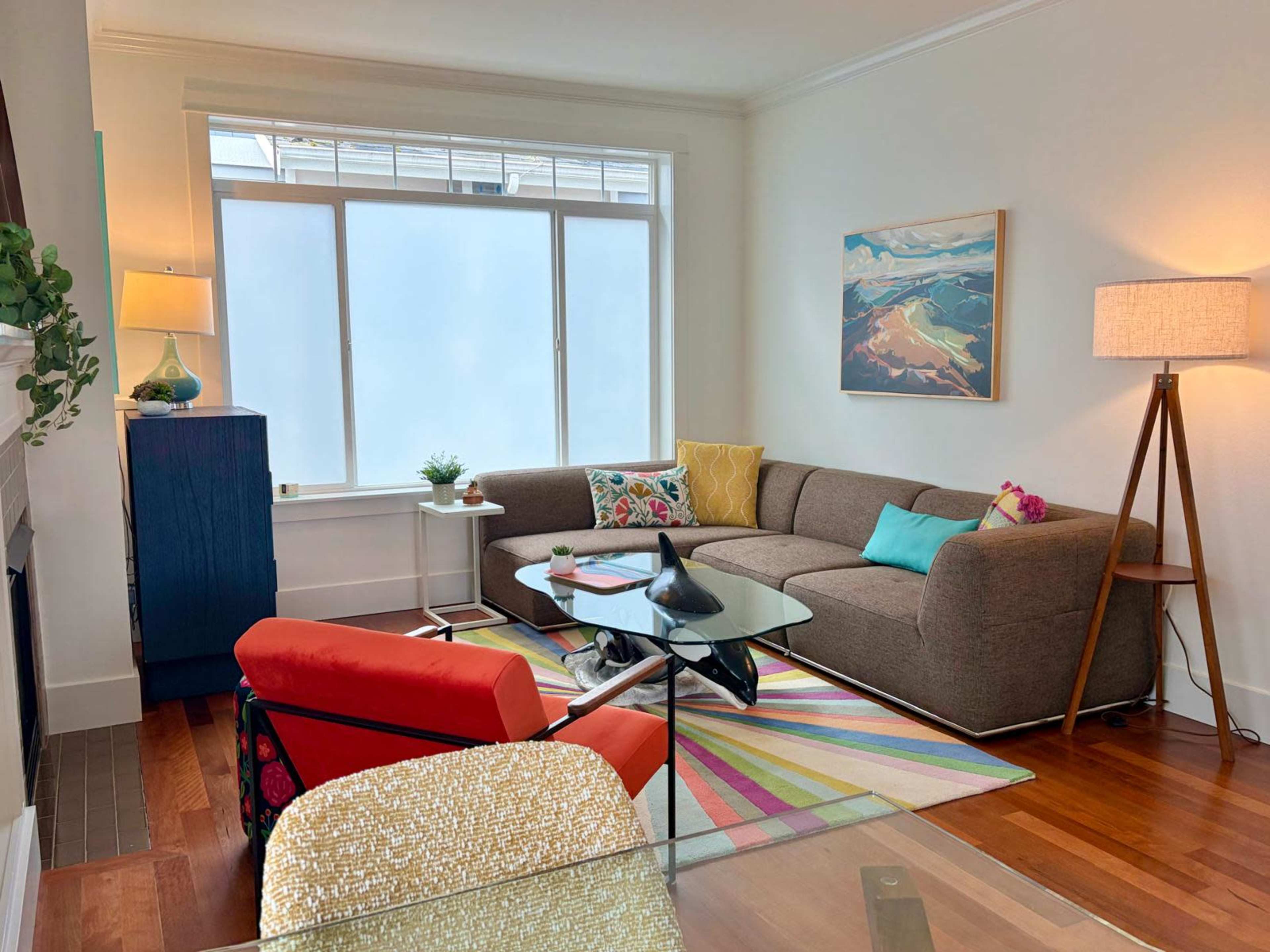 The image shows a cozy living room with a brown sectional sofa, a colorful rug, a glass coffee table, and a bright orange armchair, complemented by natural light from large windows.