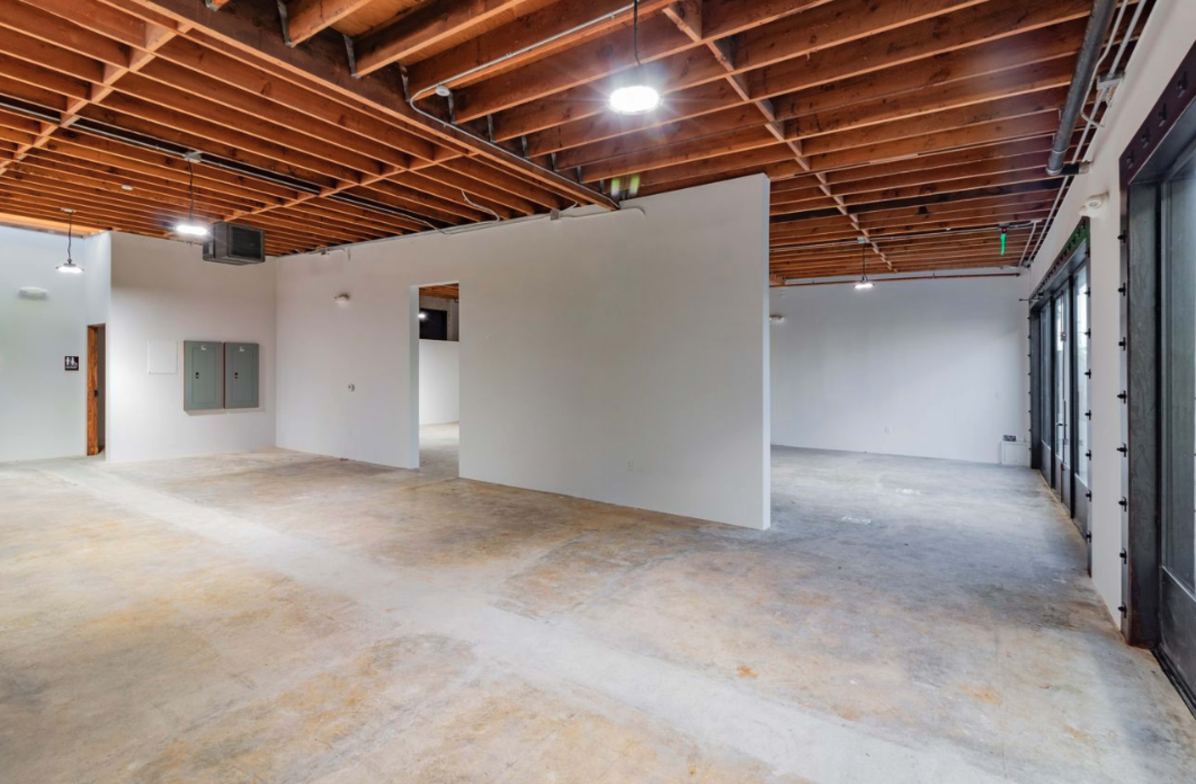 The image shows an empty interior space with exposed wooden beams, concrete floors, and white walls.