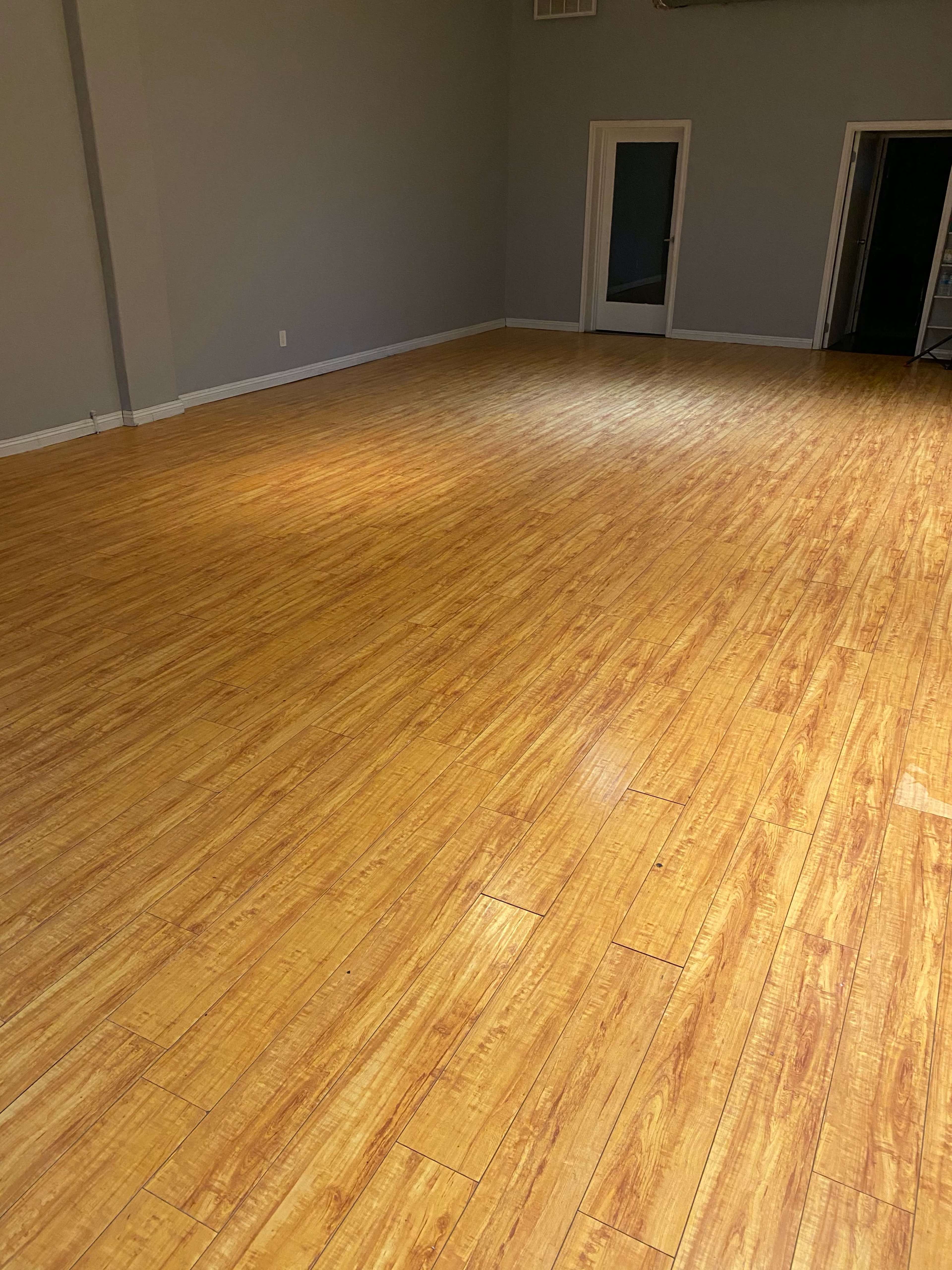 The image shows a spacious room with a polished bamboo floor and light gray walls.