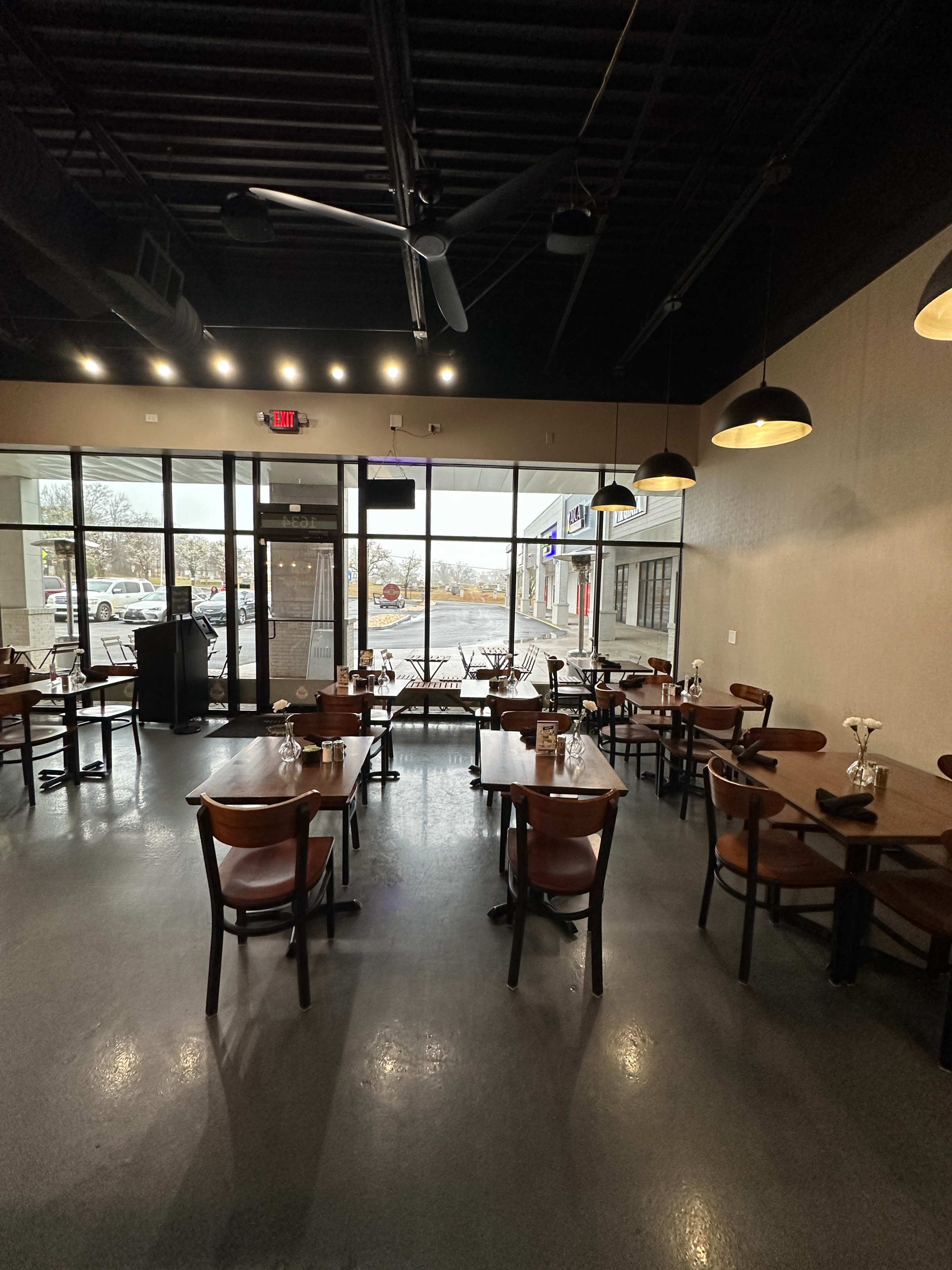 The interior of a restaurant with wooden tables and chairs, a large front window displaying the street outside, and subtle lighting fixtures hanging from the ceiling.