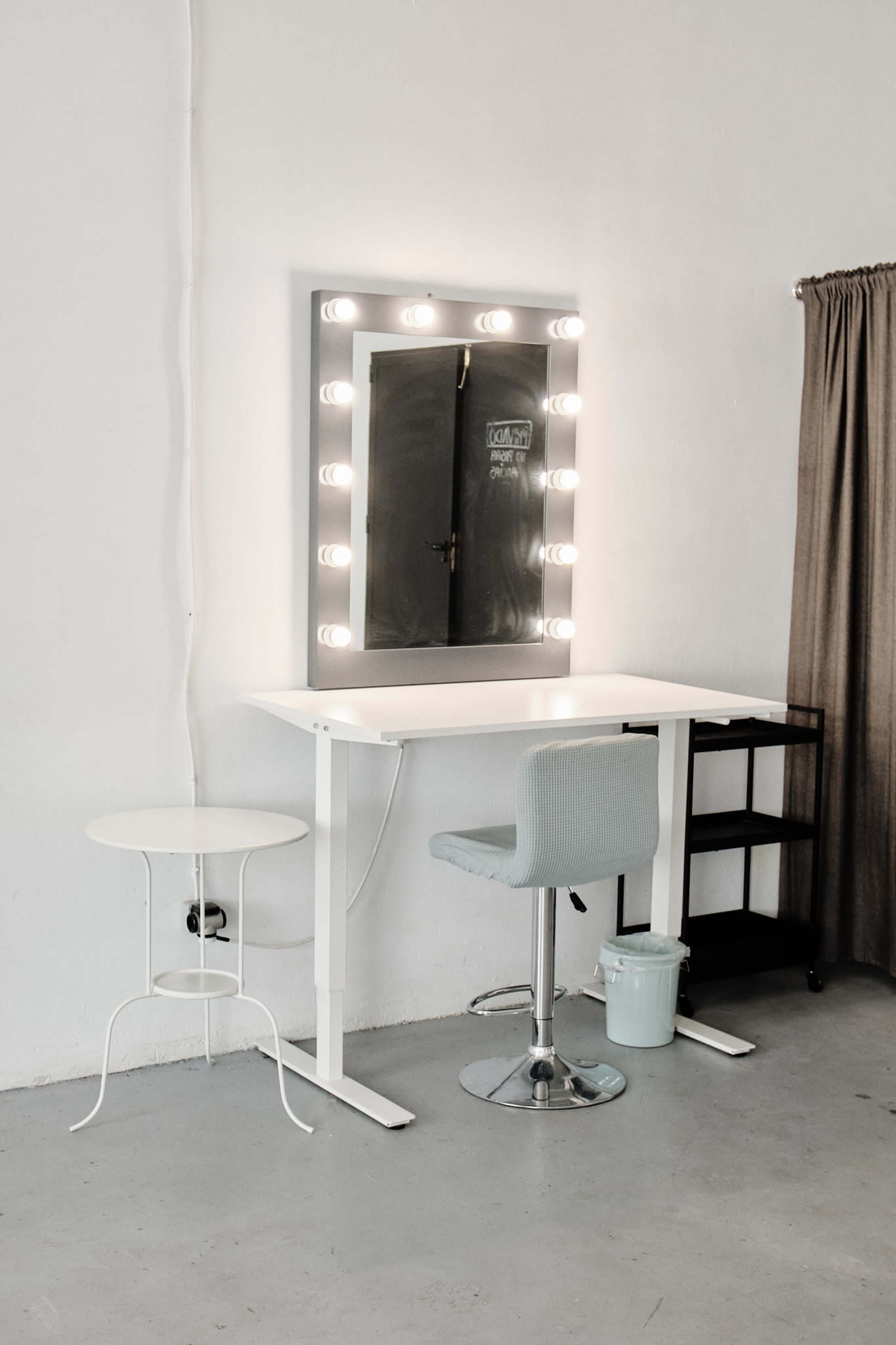 The image shows a minimalist makeup station with a lighted mirror, a white table, a blue swivel chair, and a small side table in a well-lit room.