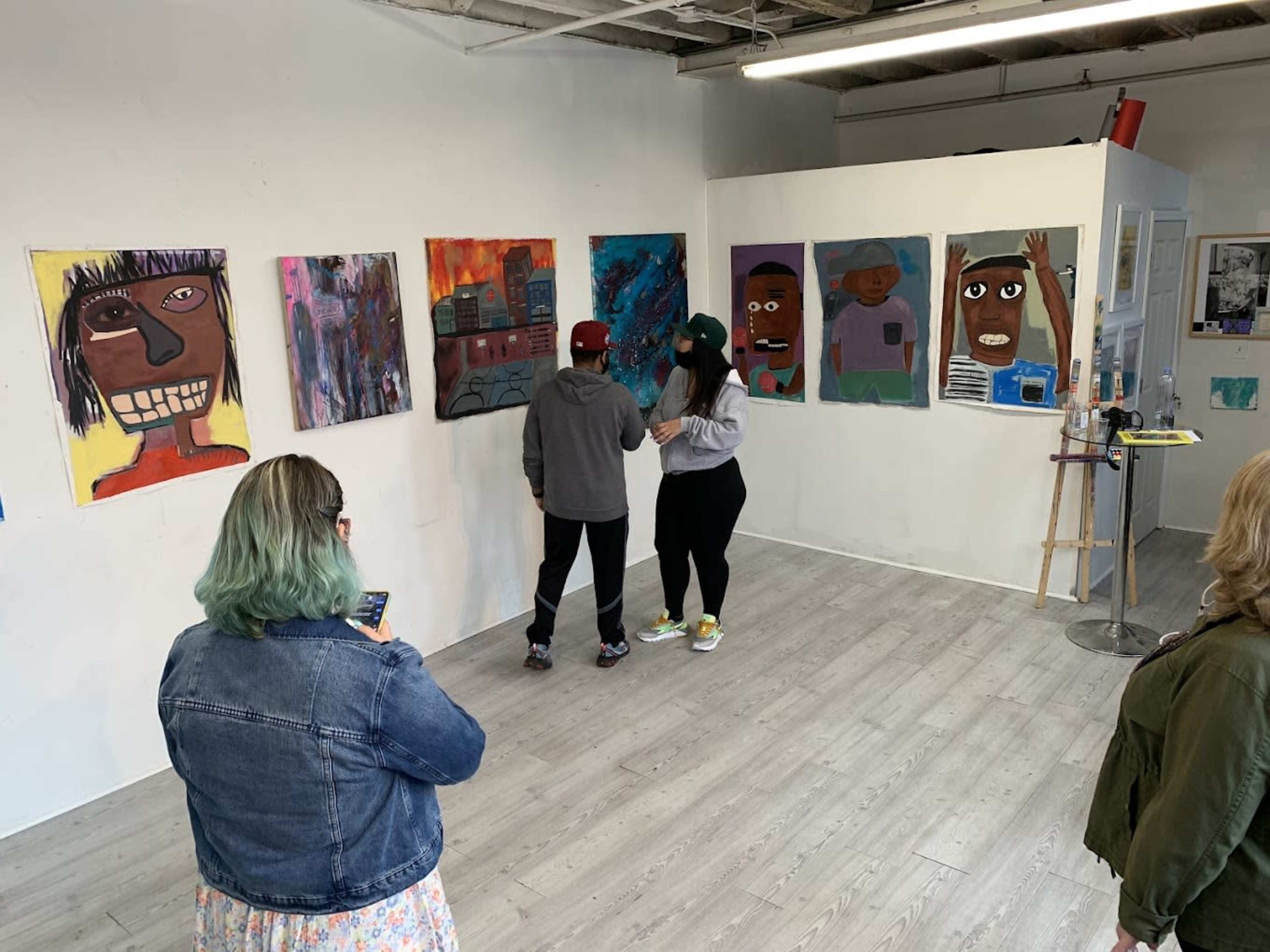Two individuals examine artwork on a gallery wall while another person stands at a distance, holding a phone and observing the scene.