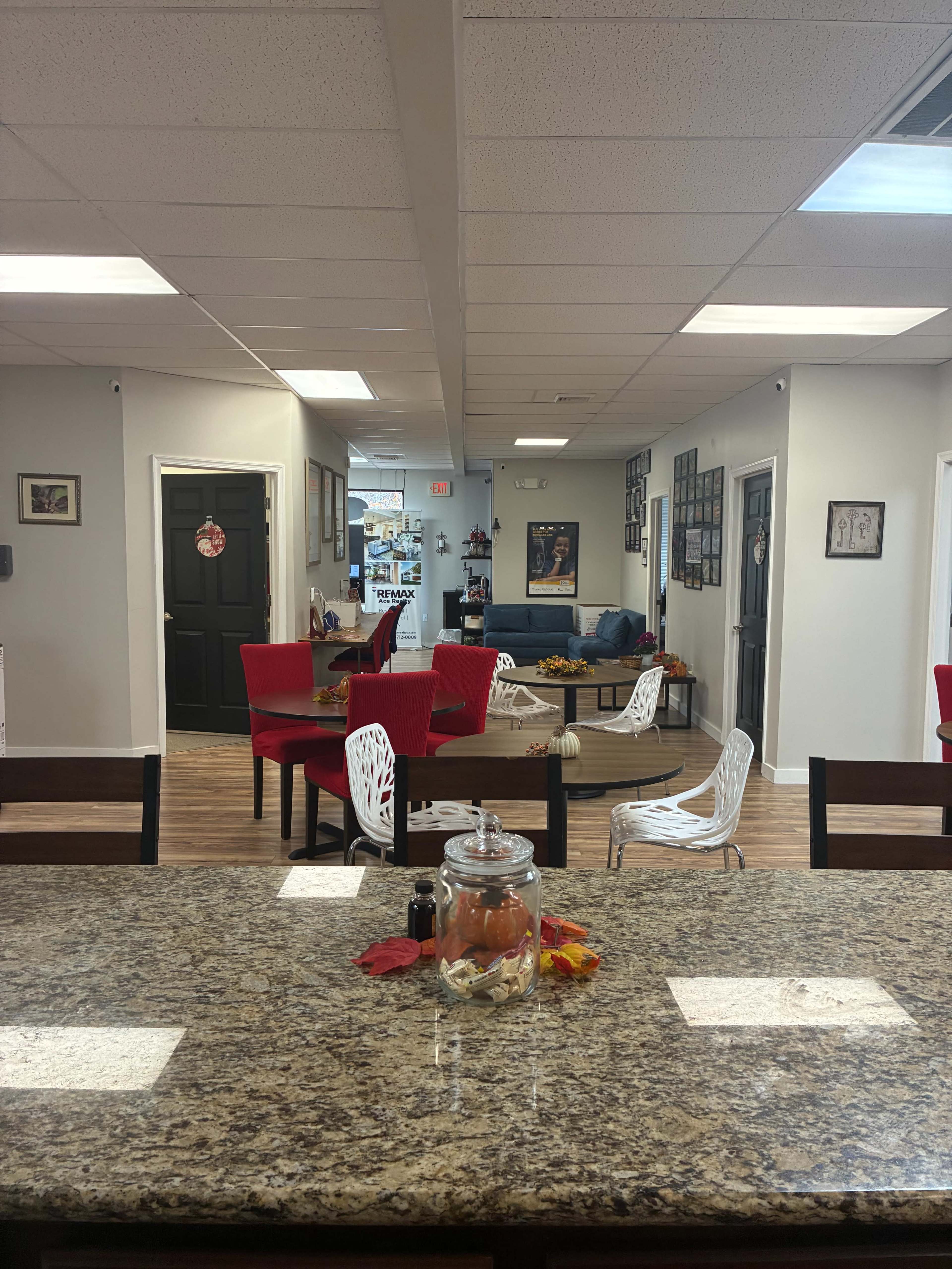 The image shows an indoor space with a kitchen area featuring a granite countertop, red chairs, and a view into a hallway with additional seating and wall decorations.