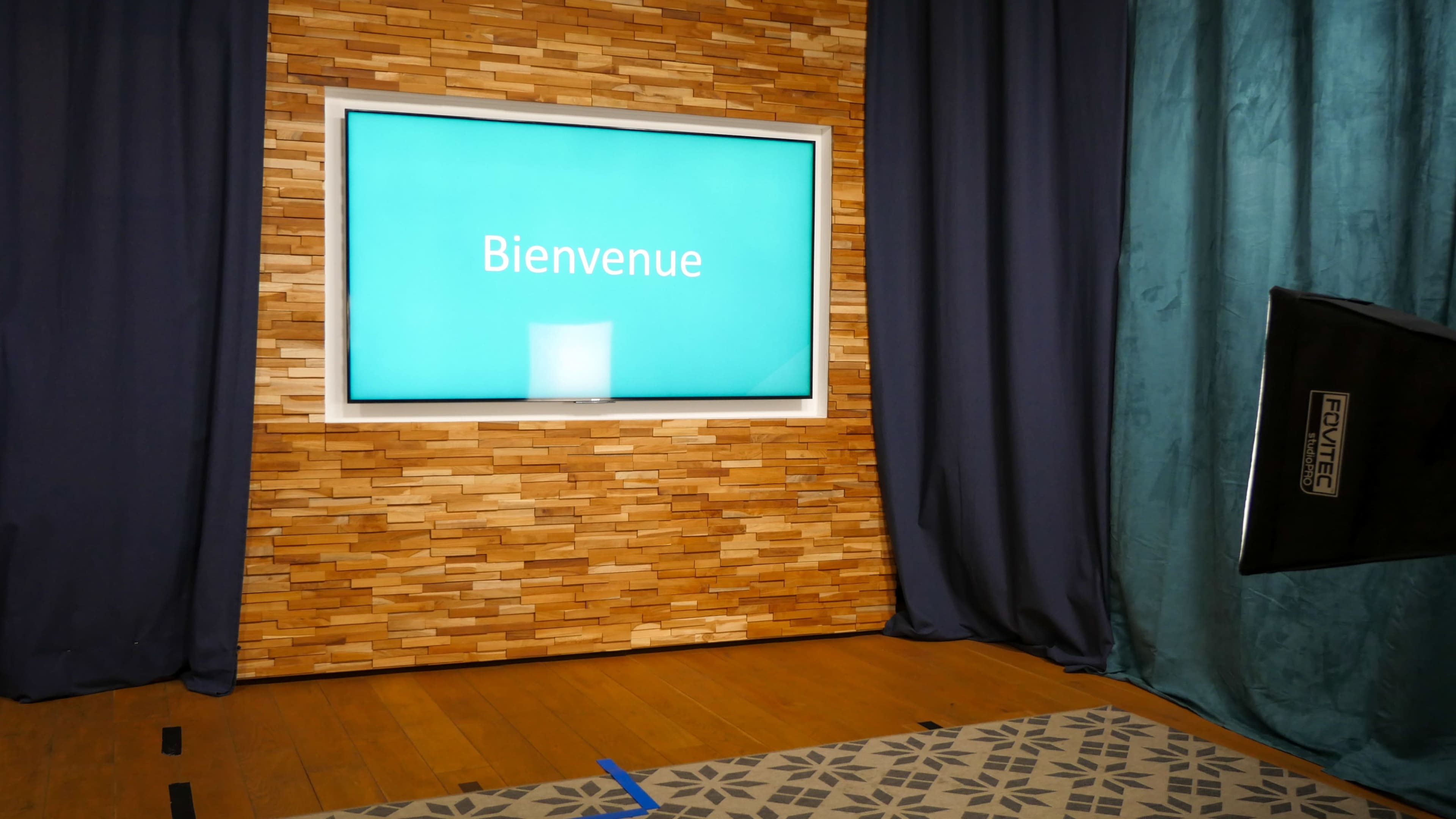 The image shows a decorated stage area with a wooden wall, a screen displaying the word "Bienvenue," and curtains on either side.