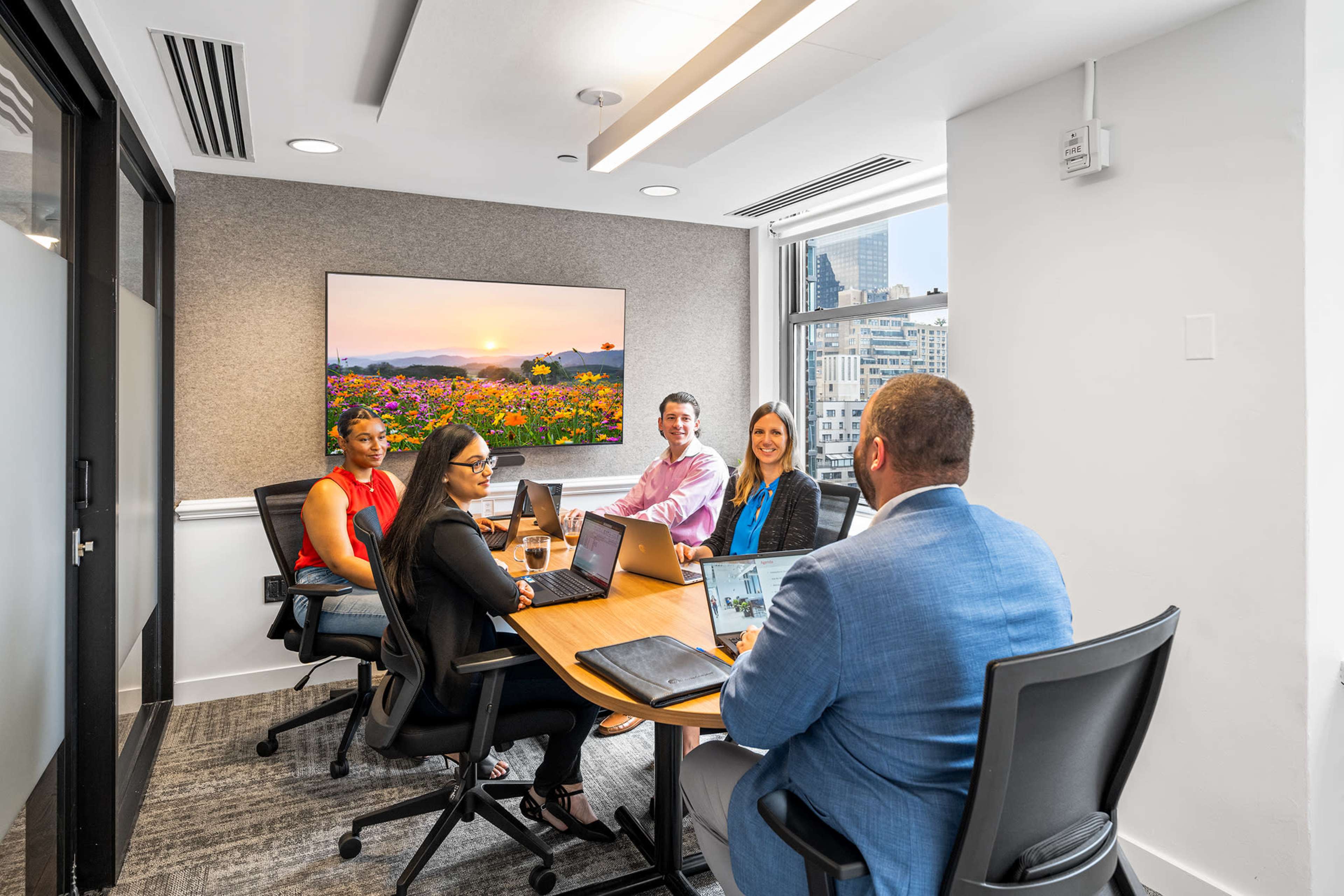 A group of five professionals is seated around a conference table in a modern meeting room, with a large screen displaying a landscape image in the background.