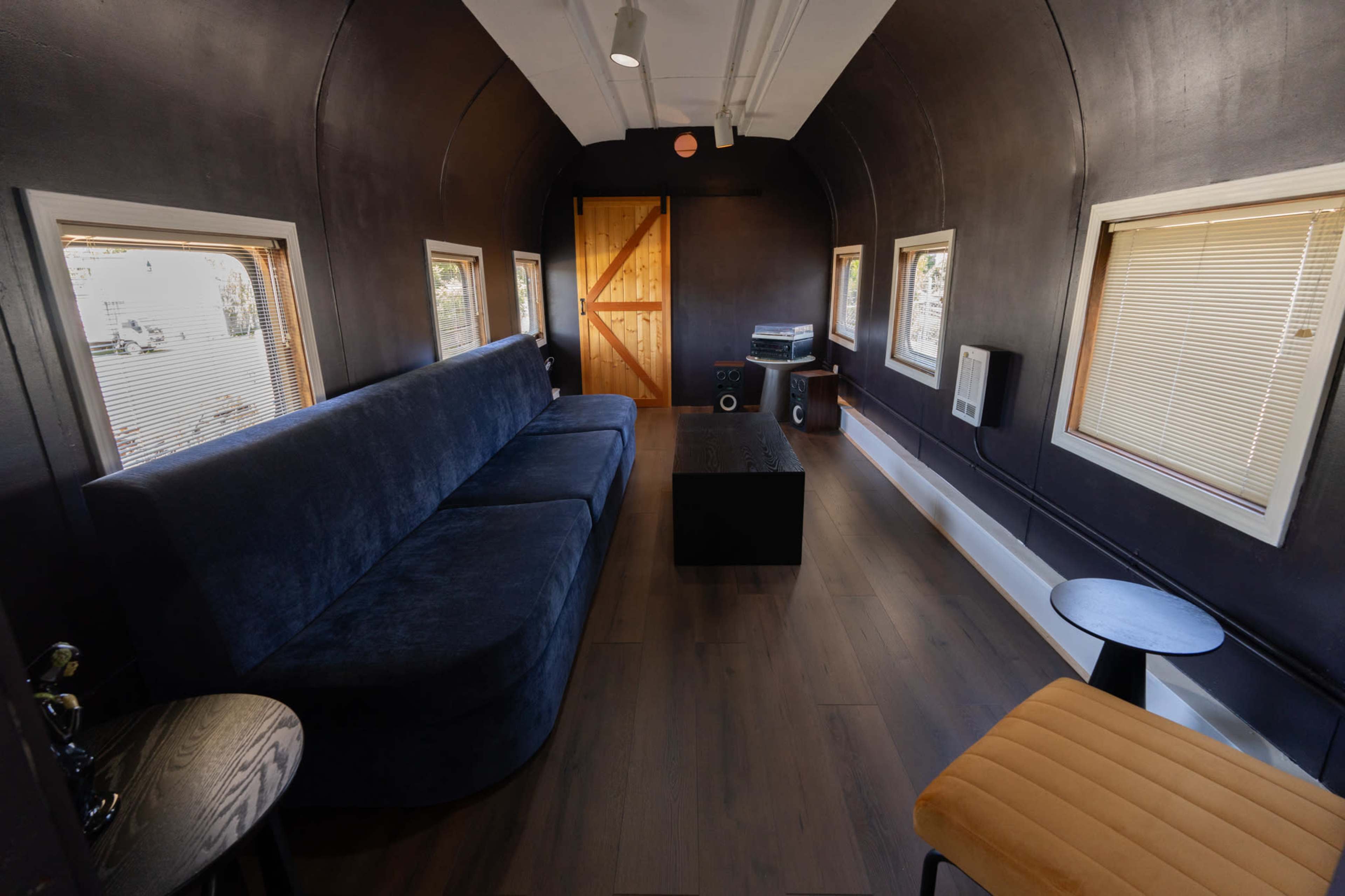 The image shows a modern, narrow interior of a room featuring a dark-colored couch, a coffee table, and a wooden door, with windows on either side allowing natural light to enter.