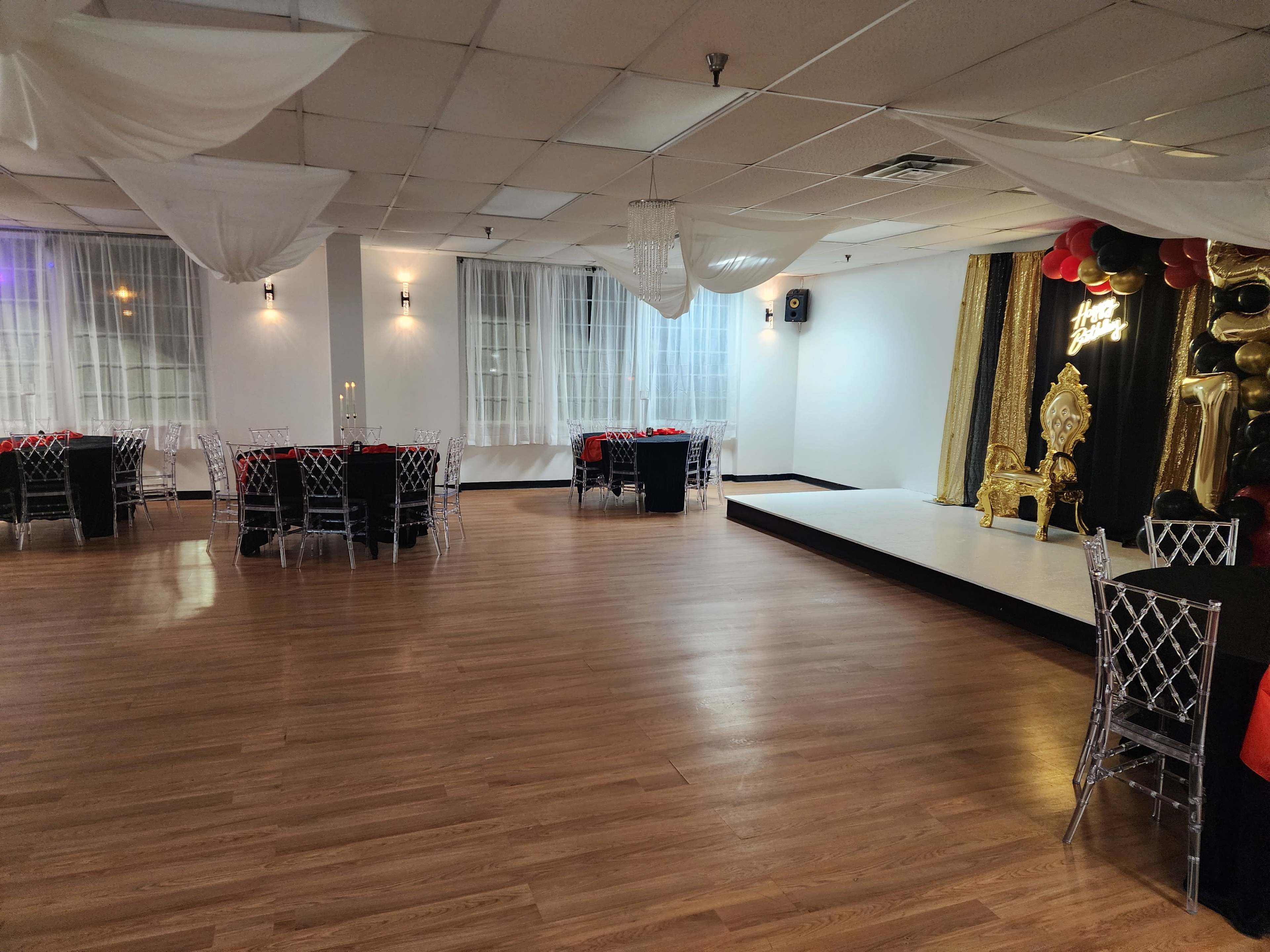 The image depicts a spacious event hall with polished wooden floors, decorated tables, and a stage area featuring a golden throne and black and red balloons.