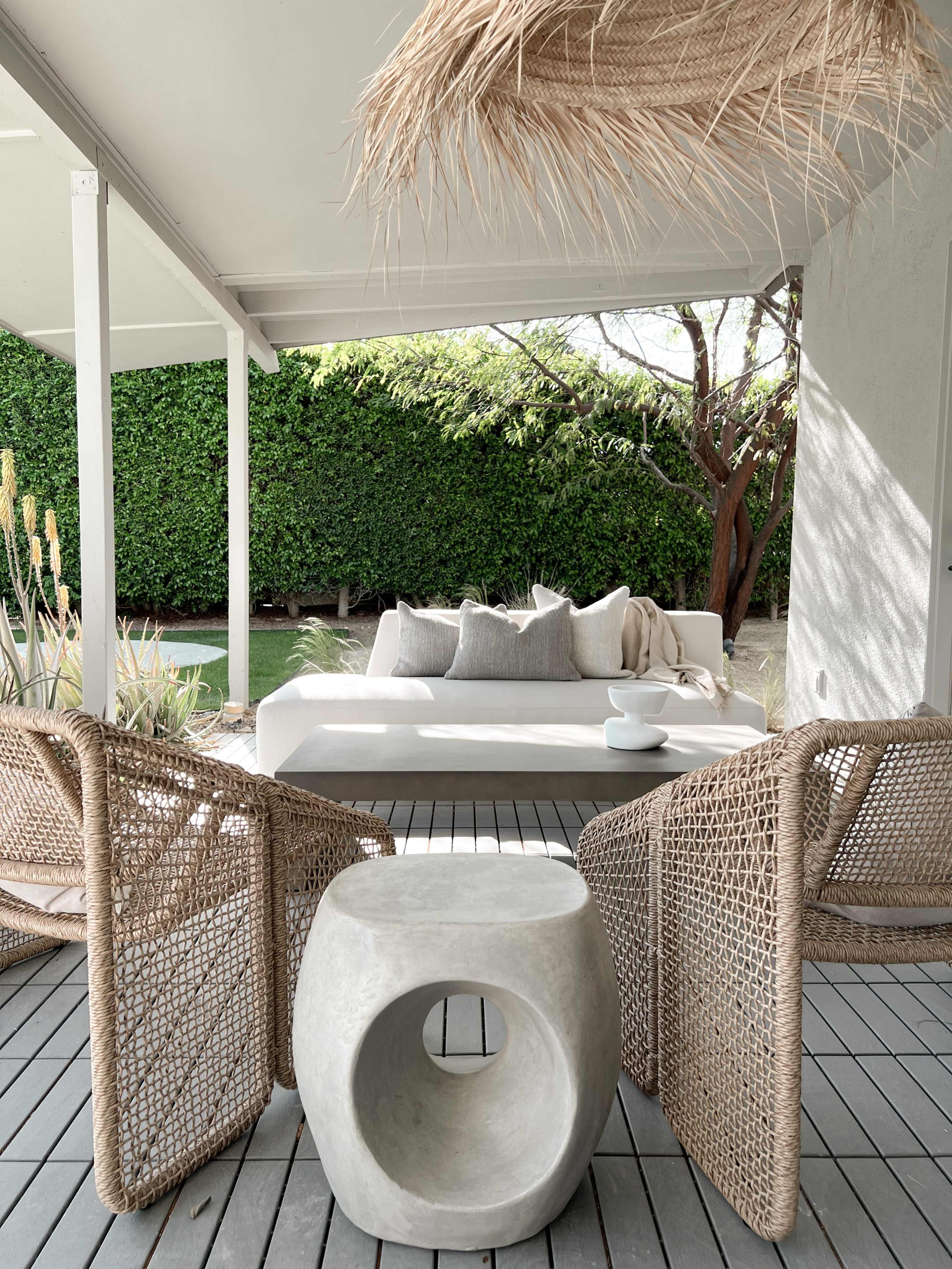 A modern outdoor seating area features a white sofa with cushions, two woven chairs, and a circular concrete side table against a backdrop of green foliage.