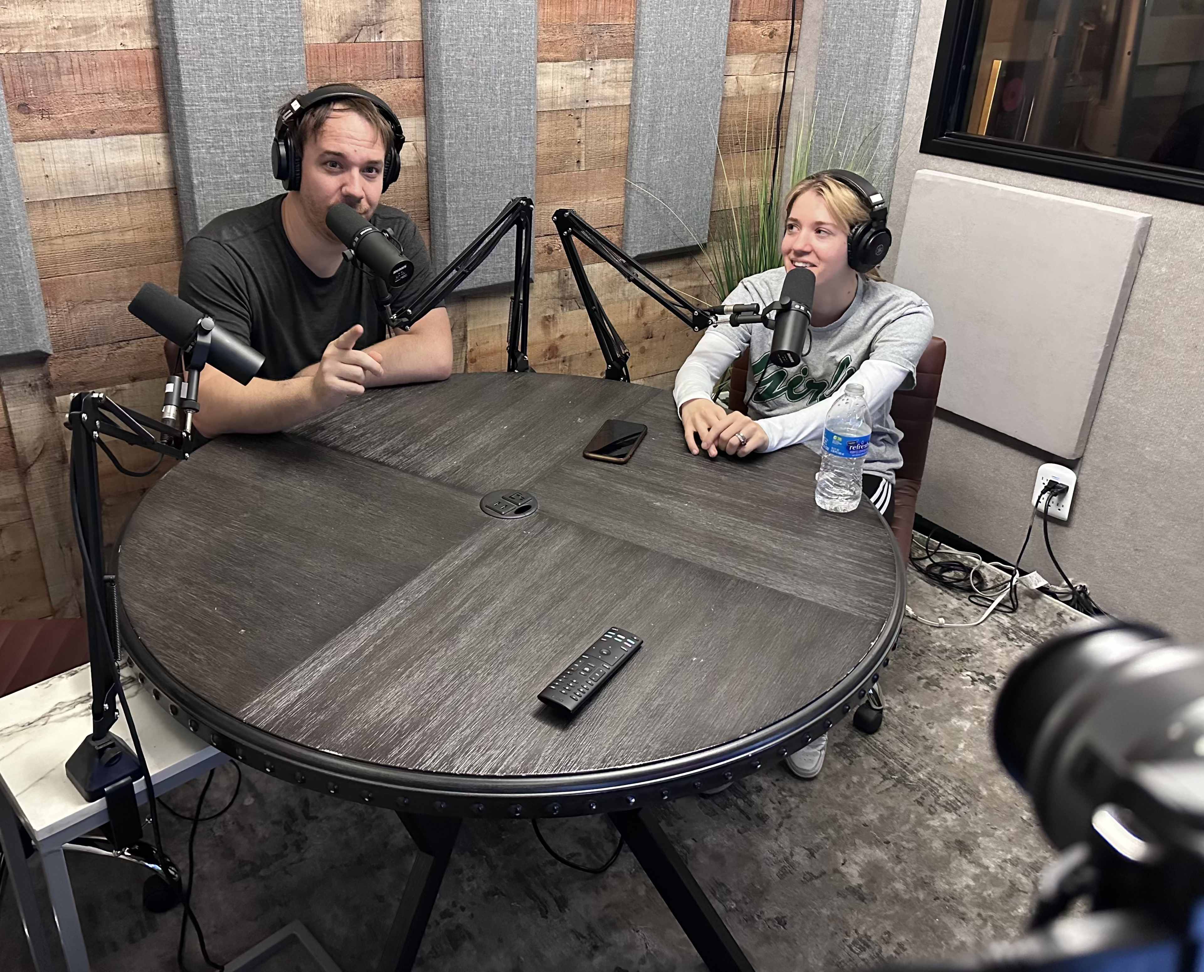 A man and a woman sit at a large round table in a recording studio, each speaking into a microphone while headphones are worn.