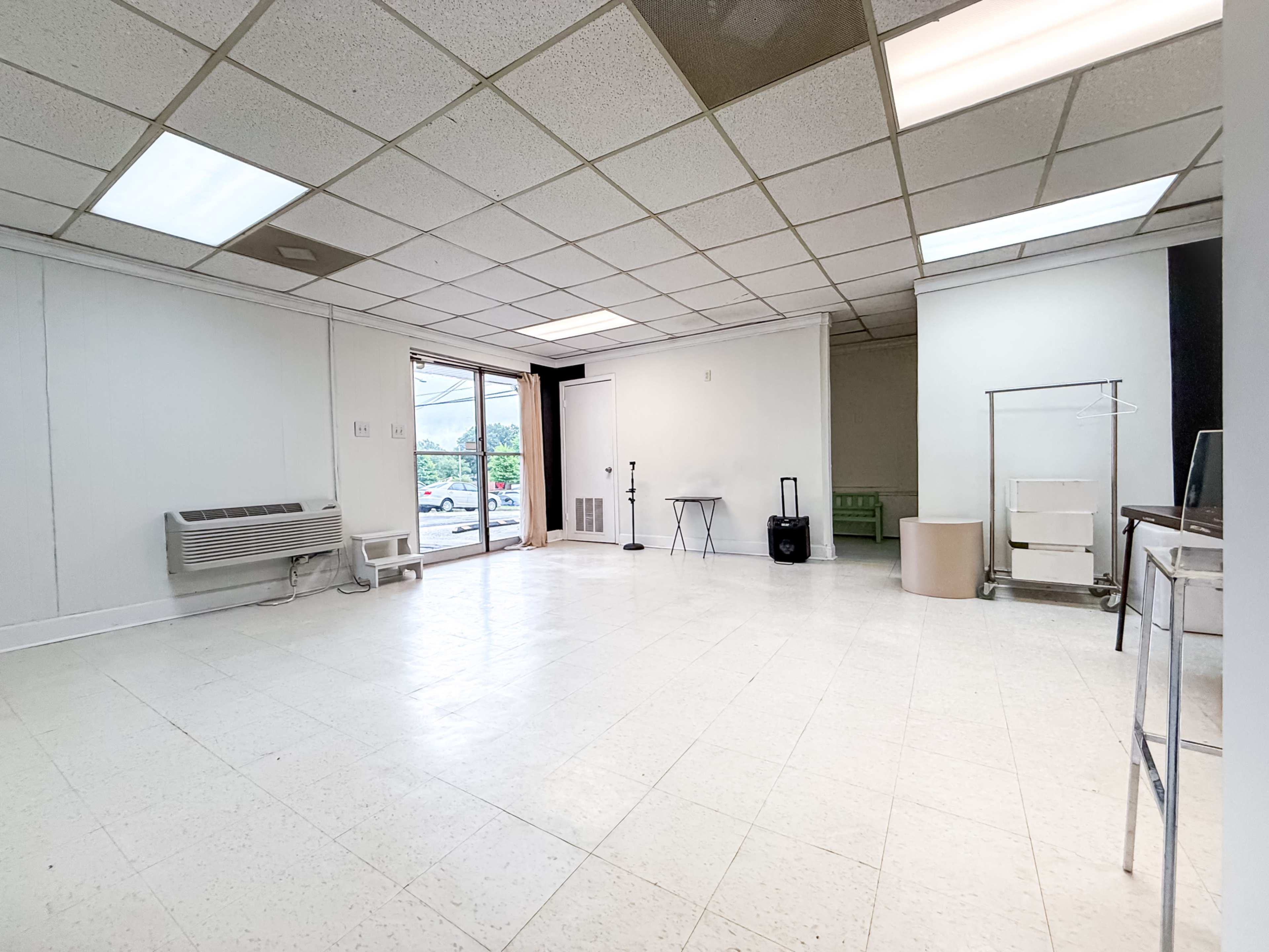 The image shows a spacious, empty room with white tiled flooring, a large window, and various minimal furnishings.