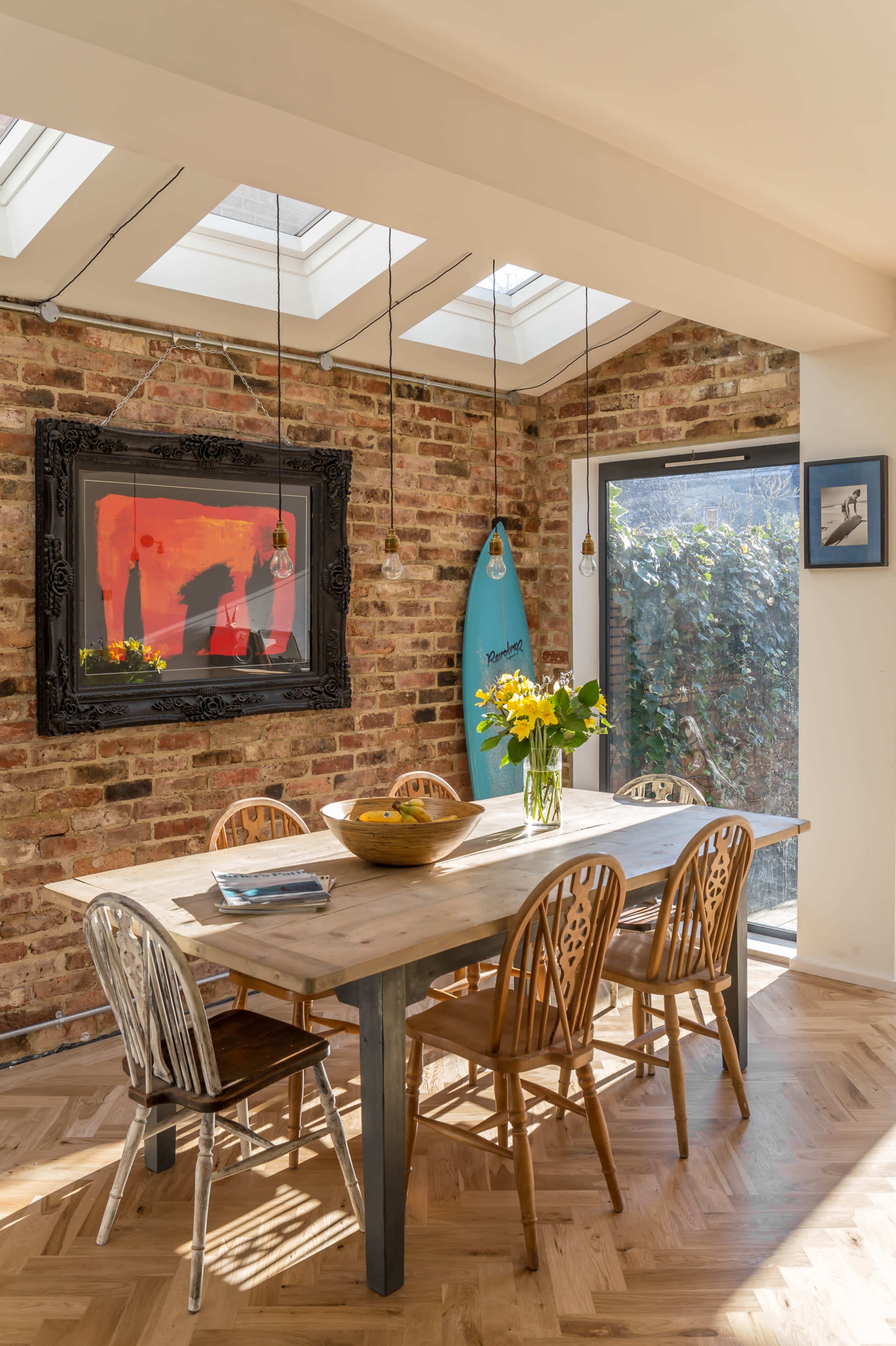A wooden dining table with eight chairs is positioned in a room with brick walls, large windows, and a surfboard hanging on the wall.