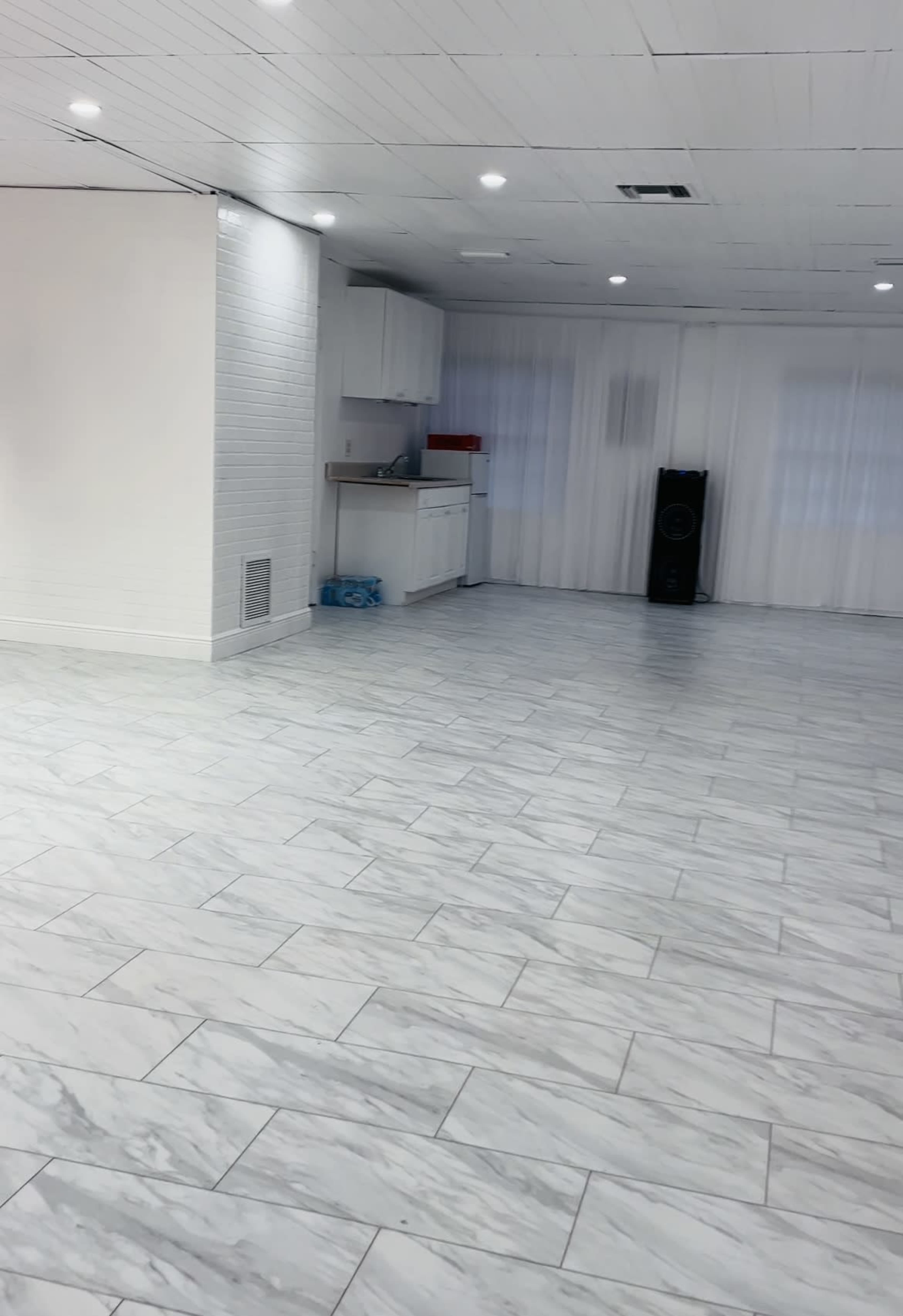 A large, empty room with white tiled flooring, a kitchenette in the corner, and large windows covered with sheer curtains.