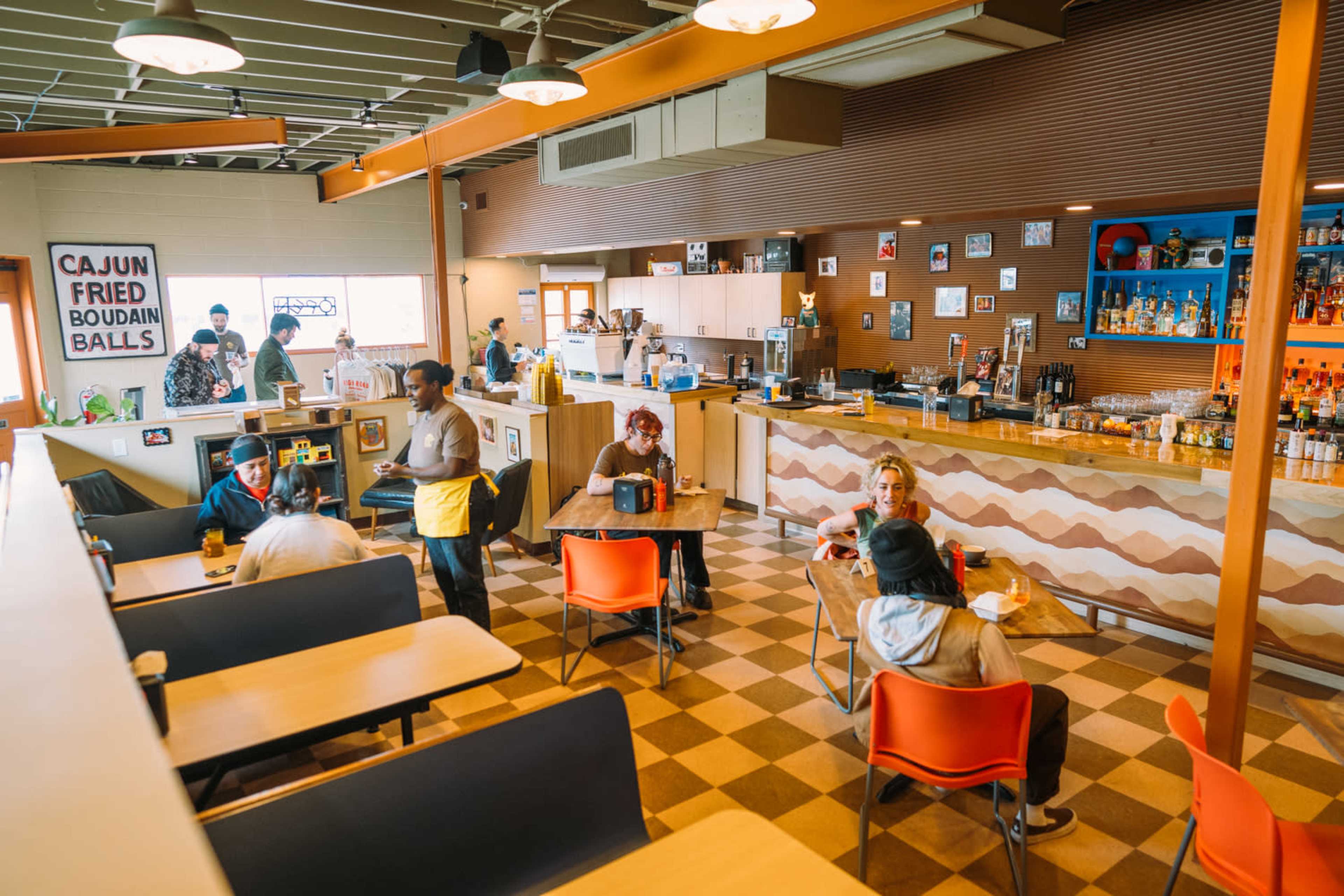 The image shows a bustling restaurant interior with patrons seated at tables and a counter displaying a variety of food and drink options.