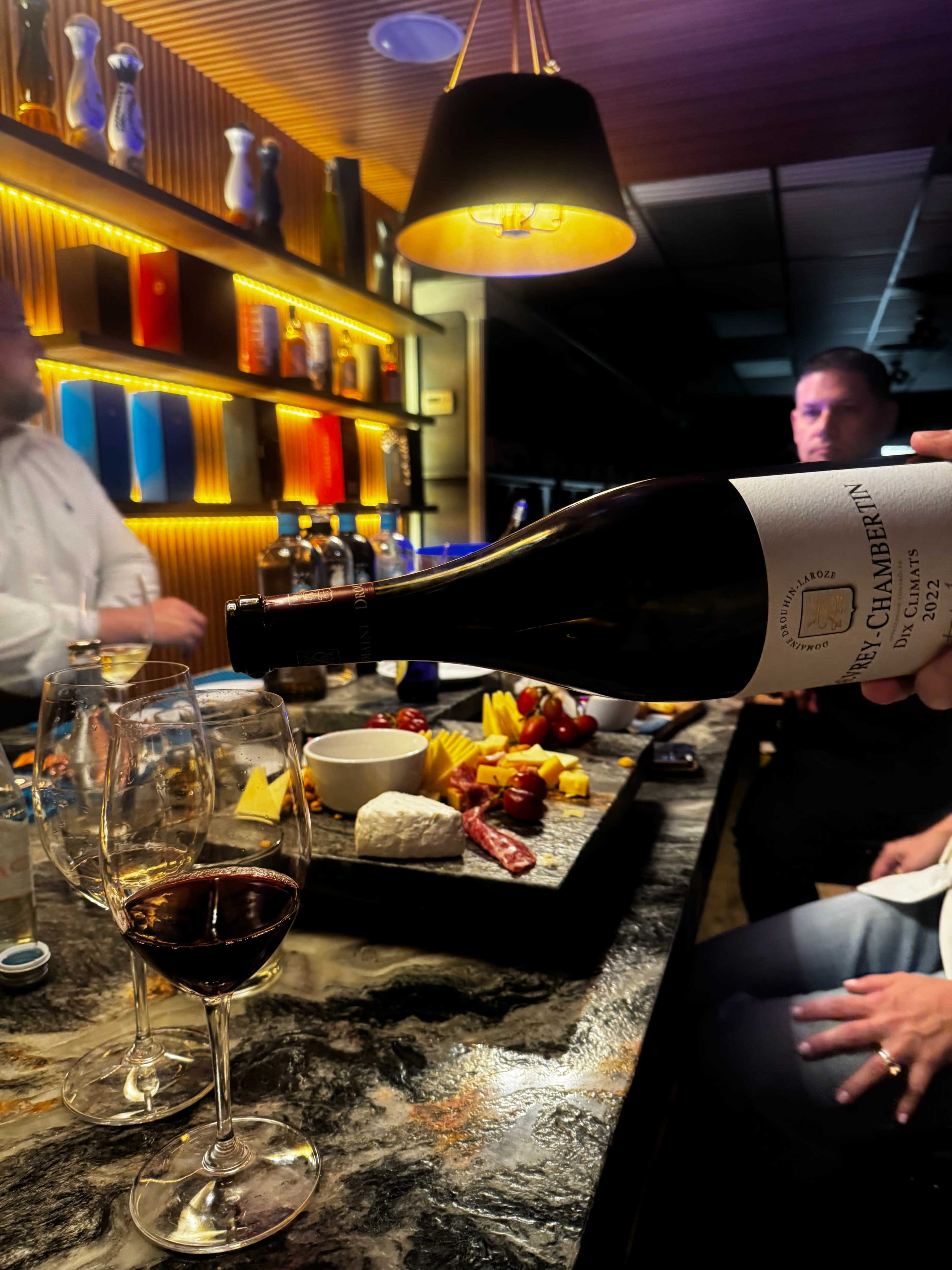 A hand pours red wine from a bottle into glasses on a bar top adorned with a charcuterie board and colorful bottles in the background.