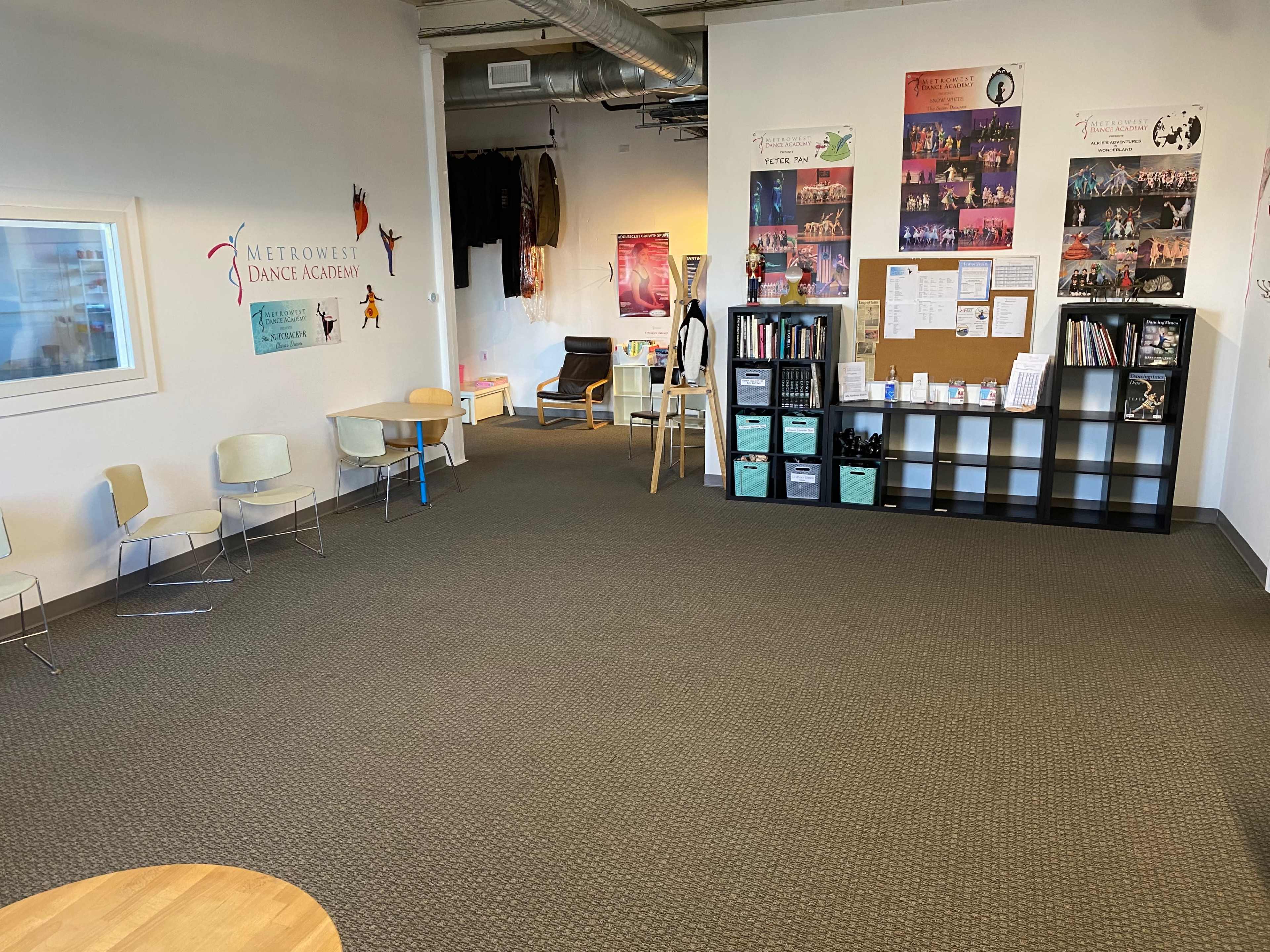 The image shows an empty dance studio with a seating area, posters on the walls, and storage shelves filled with supplies.