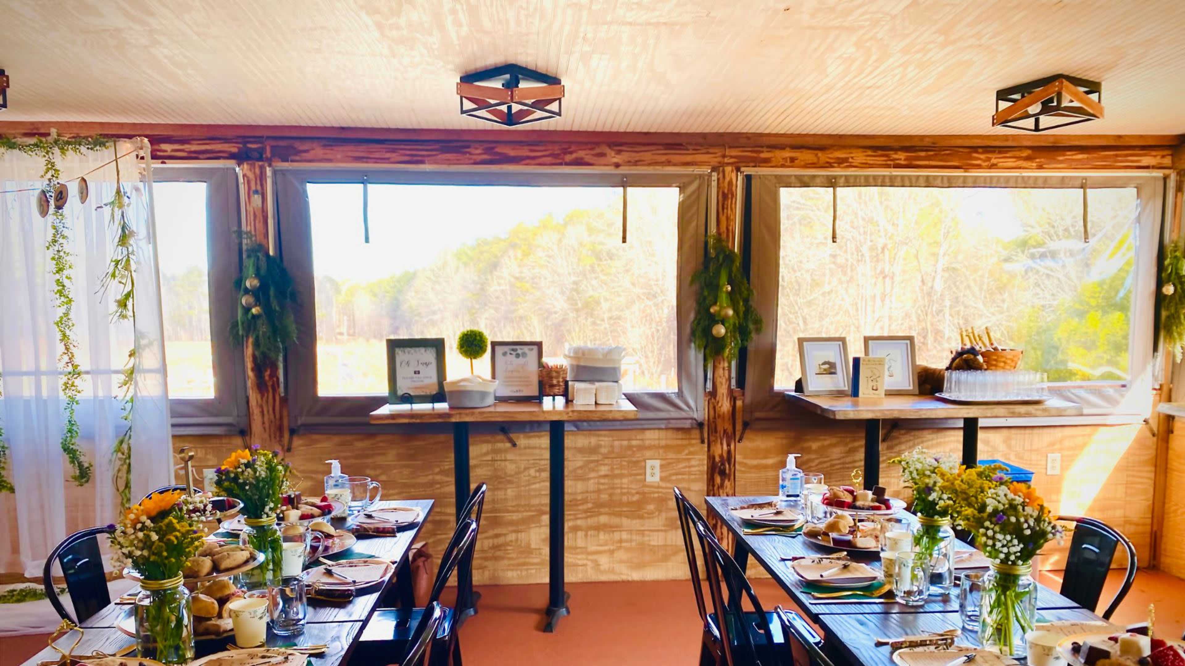 A well-set dining area with tables arranged for a meal, featuring flowers, dishes, and decor, surrounded by large windows looking out at a natural landscape.
