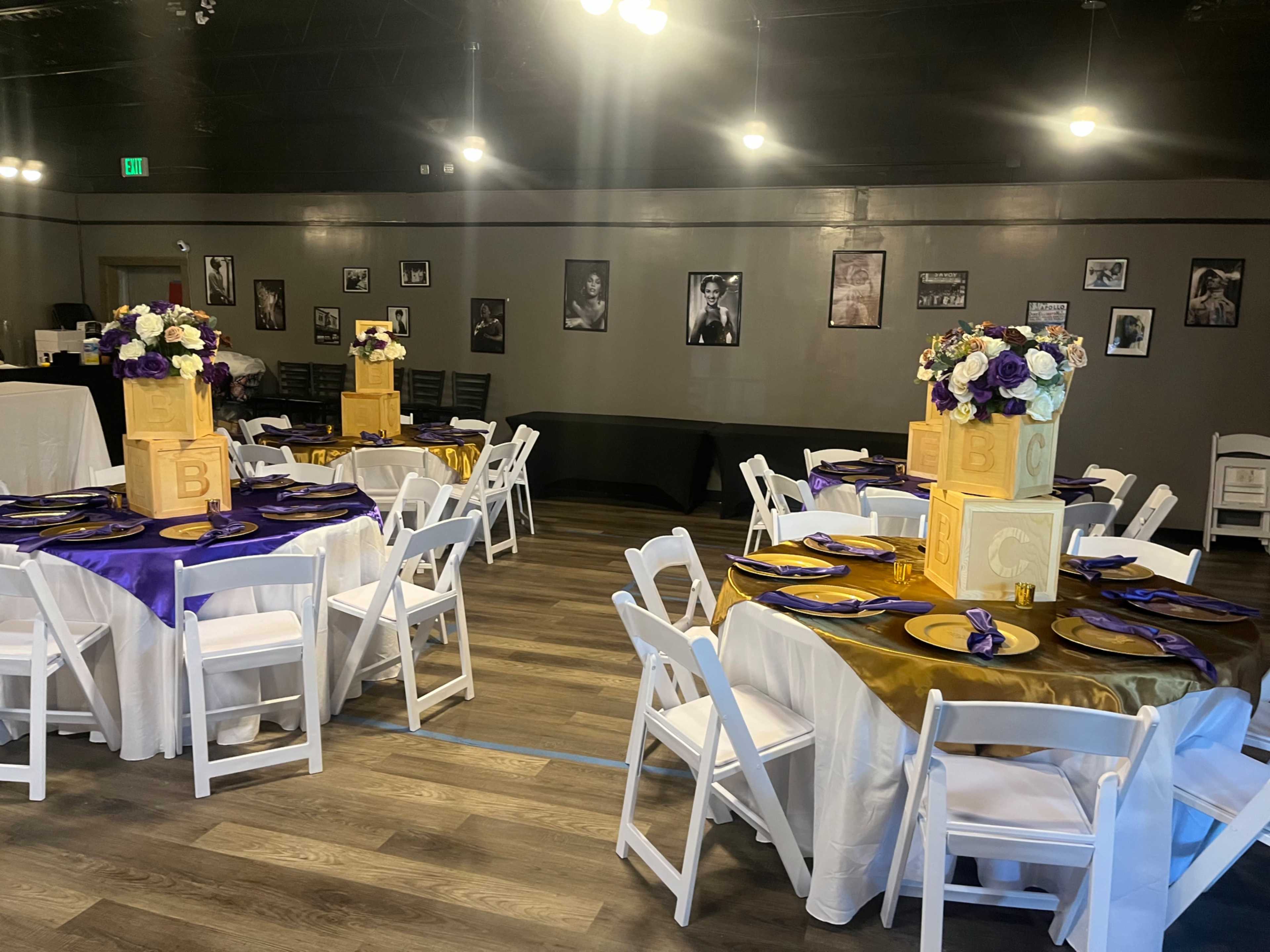 A banquet hall with several round tables decorated with white tablecloths, gold and purple accents, and floral centerpieces, surrounded by chairs and framed photographs on the walls.