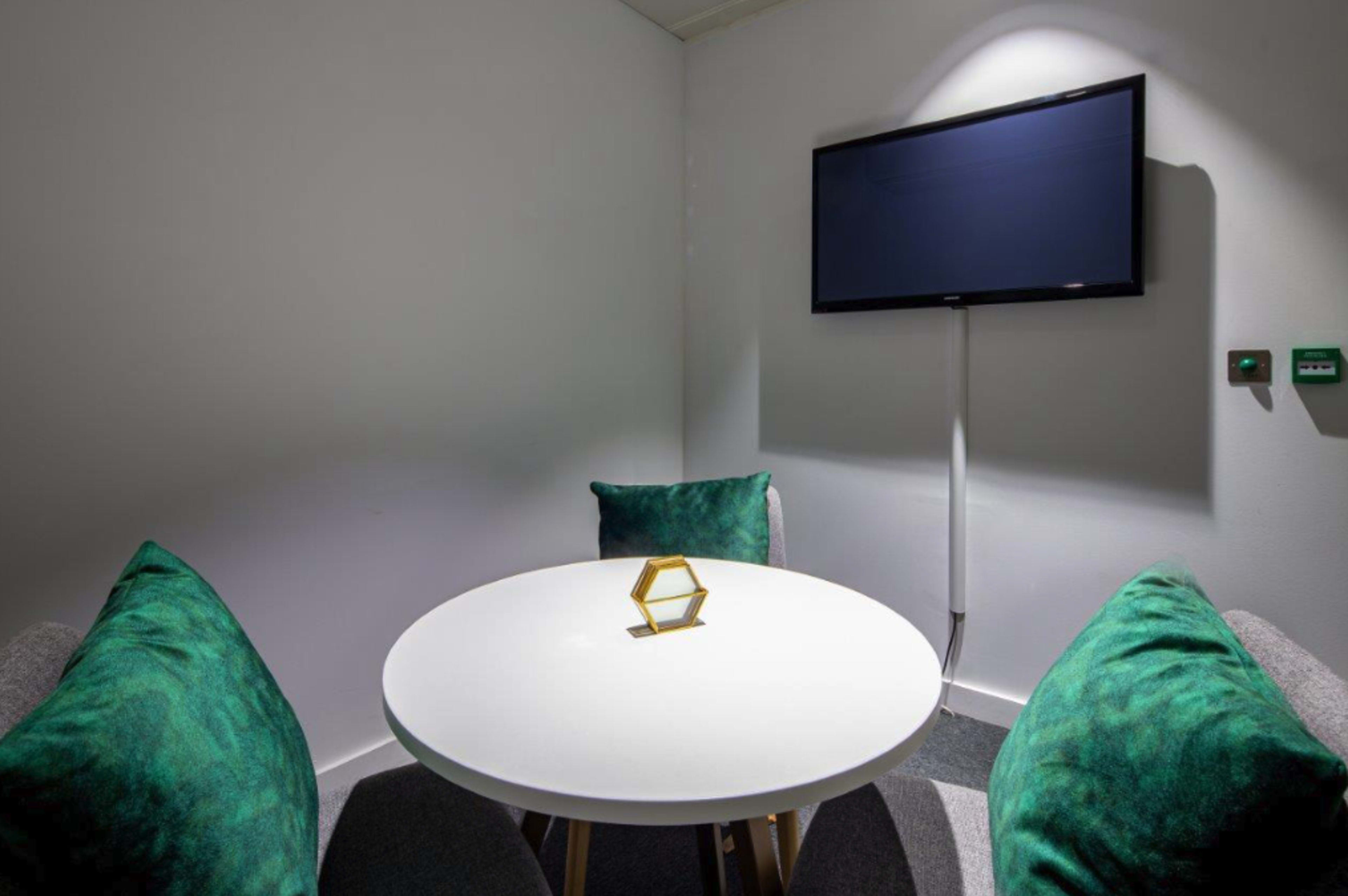 A small meeting room features a round white table, two green-cushioned chairs, and a mounted flat-screen TV on the wall.
