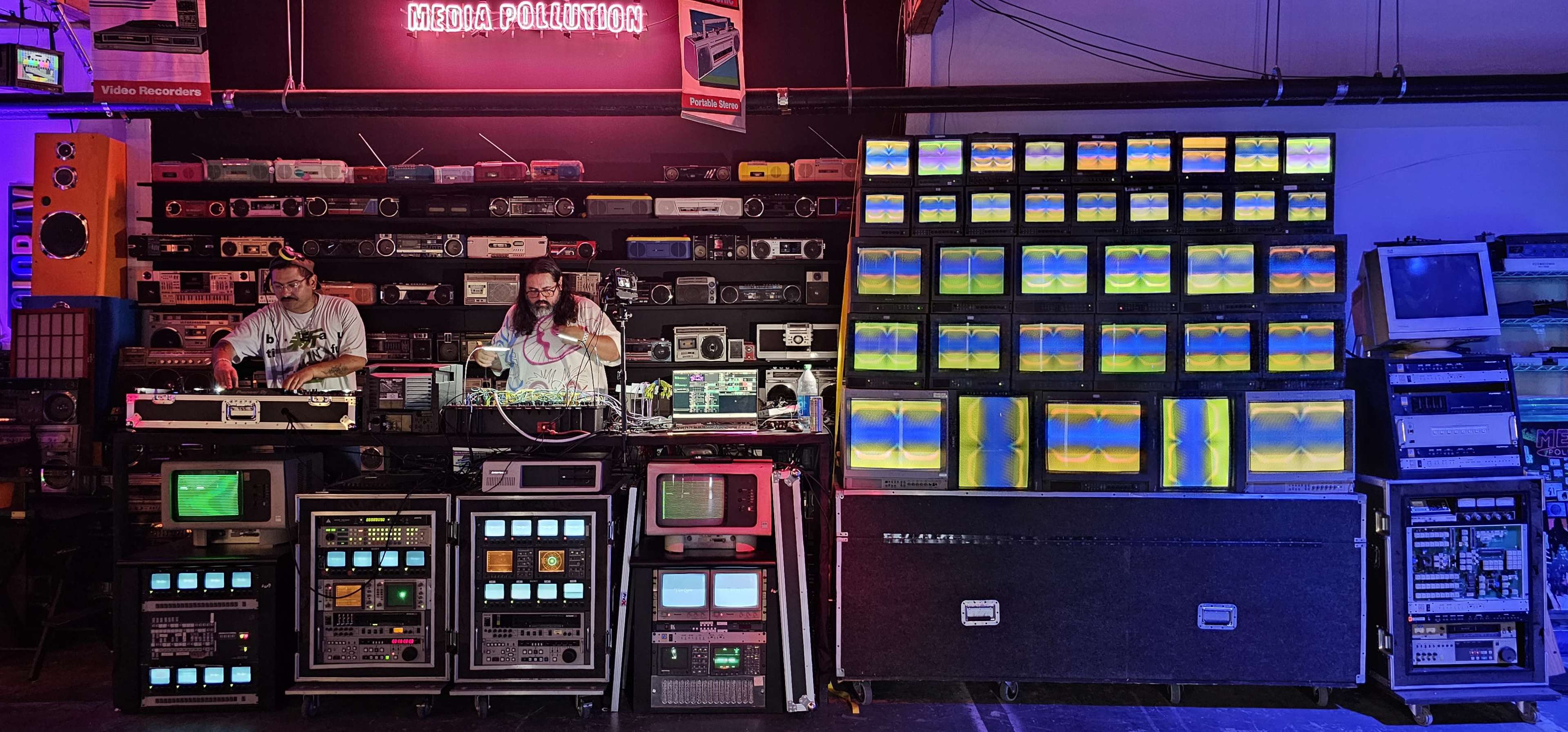 Two men are performing on a stage surrounded by an array of vintage televisions, tape players, and visual displays.