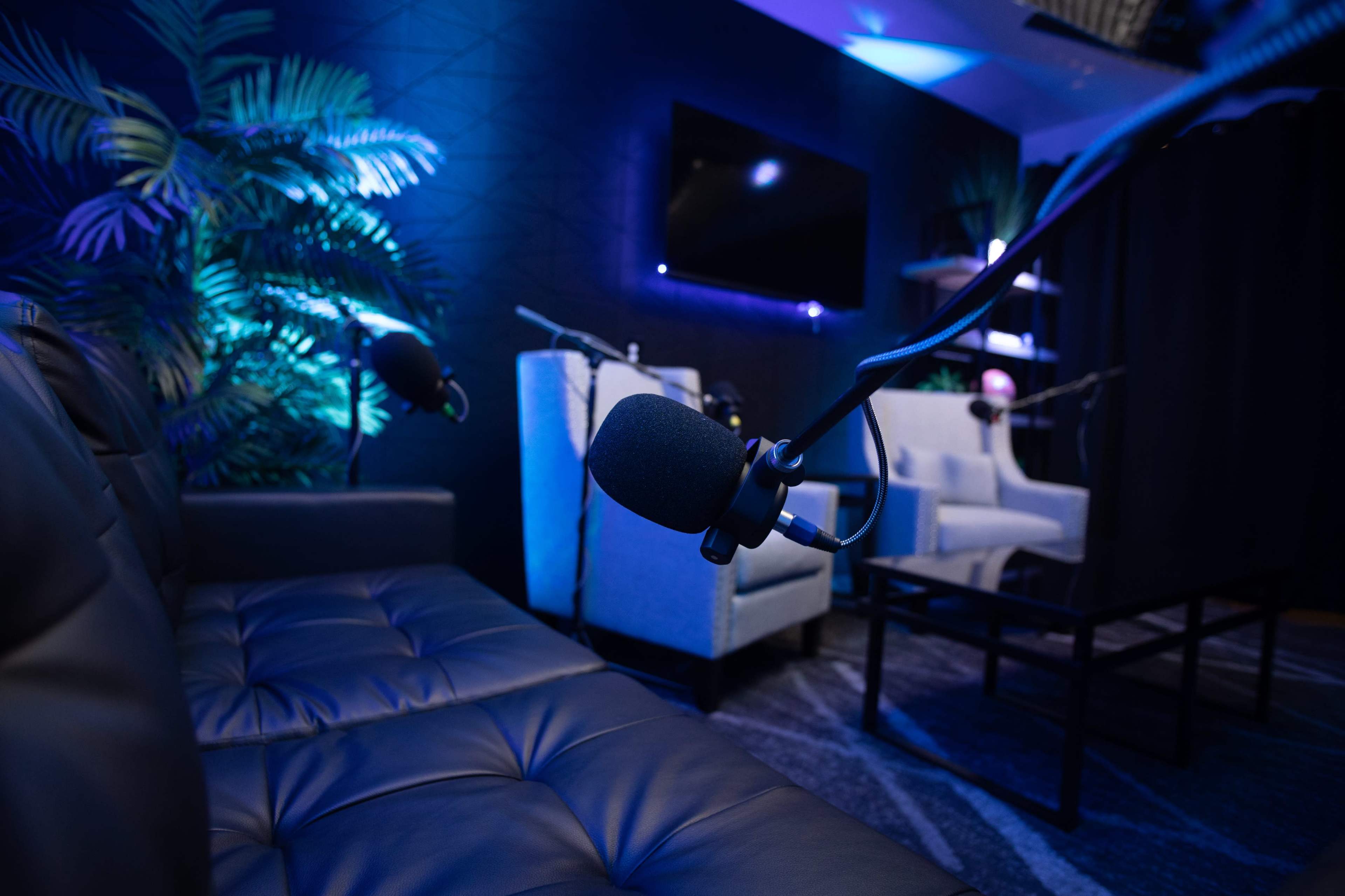 A modern recording studio with a black leather couch, two upholstered chairs, and microphones positioned on stands.